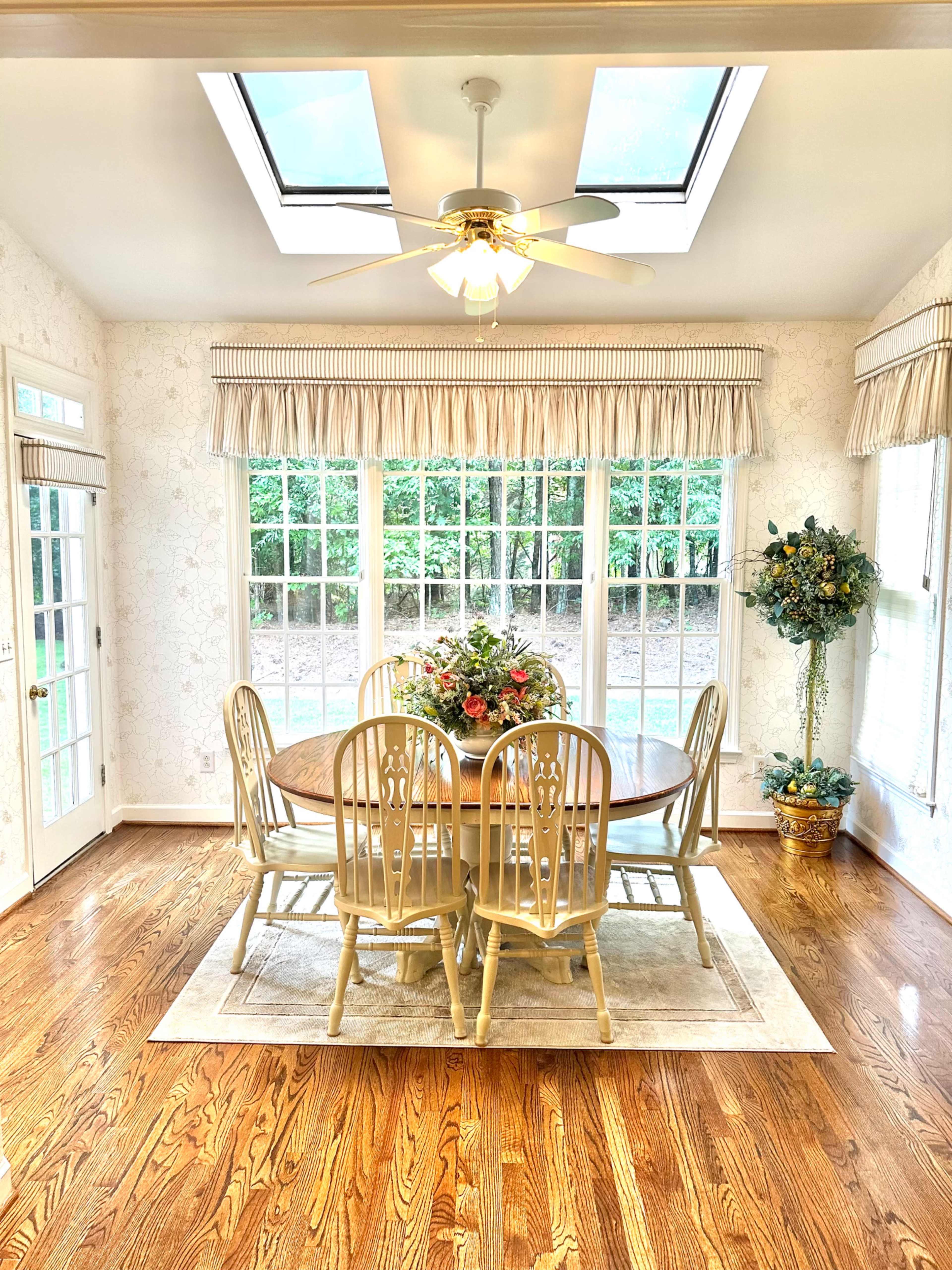 The image shows a well-lit dining area with a round wooden table surrounded by six light-colored chairs, a ceiling fan, and large windows letting in natural light.
