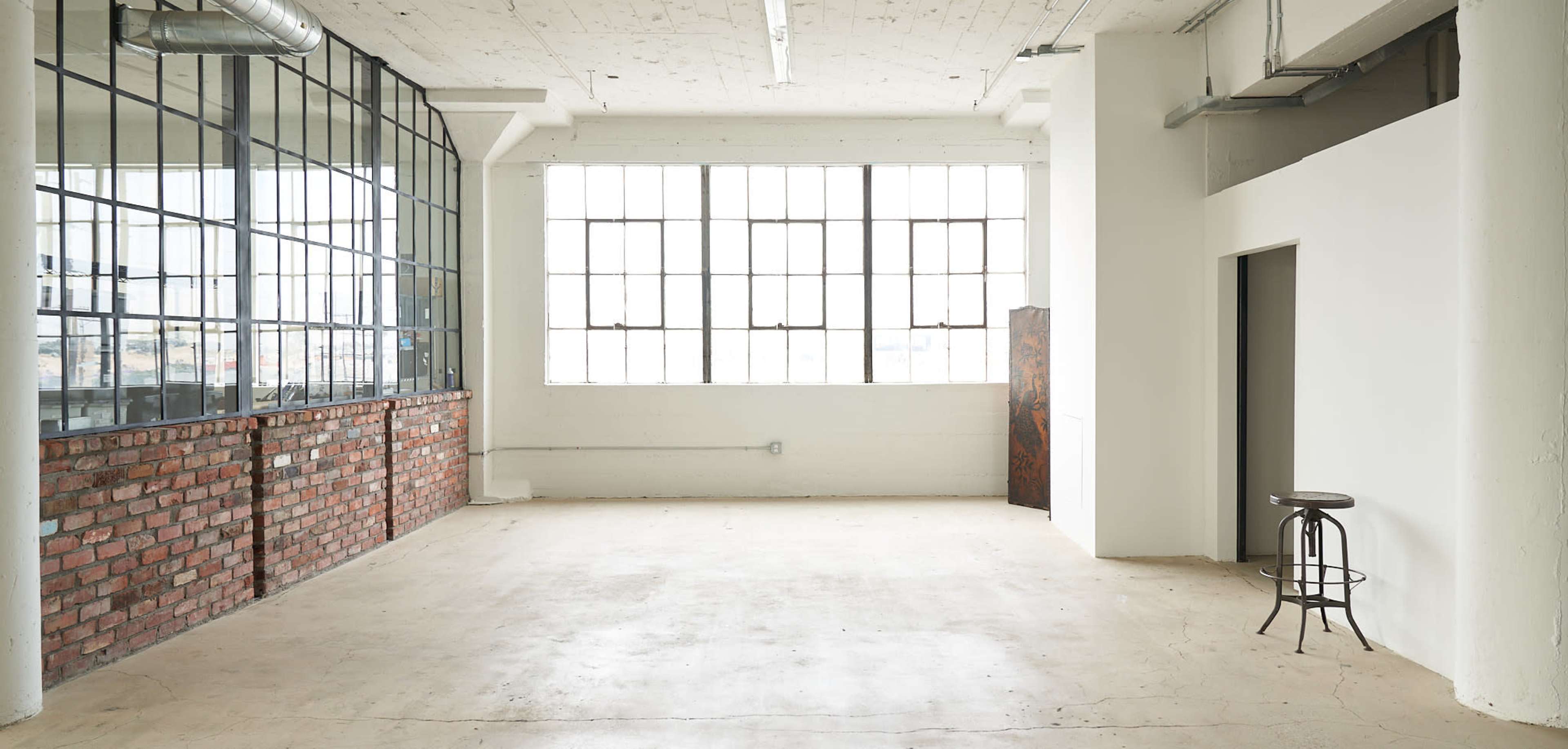 An empty interior space with large windows, exposed brick walls, and minimalistic furnishings.