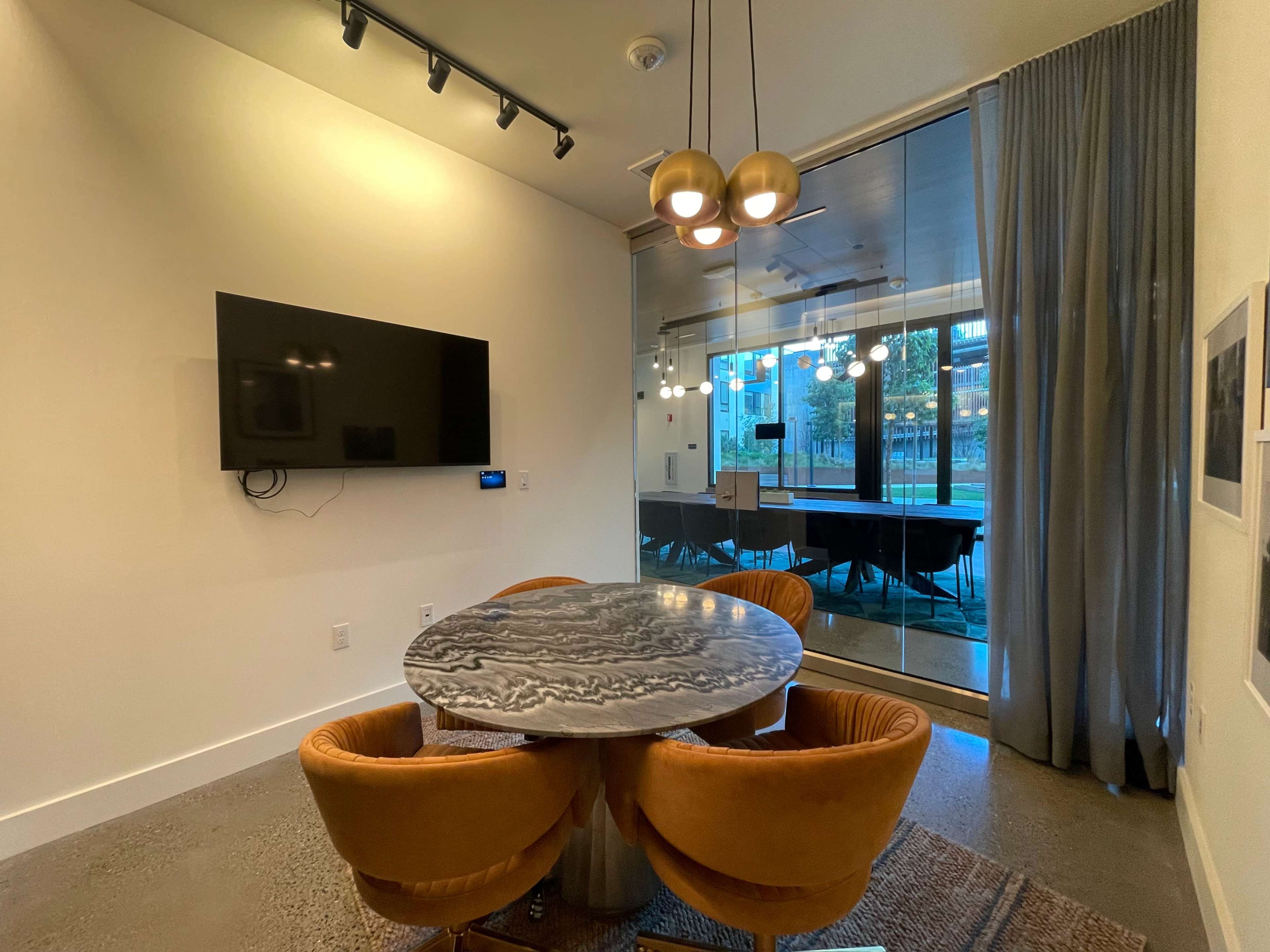 A modern meeting room features a round table with orange chairs, a large television mounted on the wall, and glass doors leading to an outdoor area.
