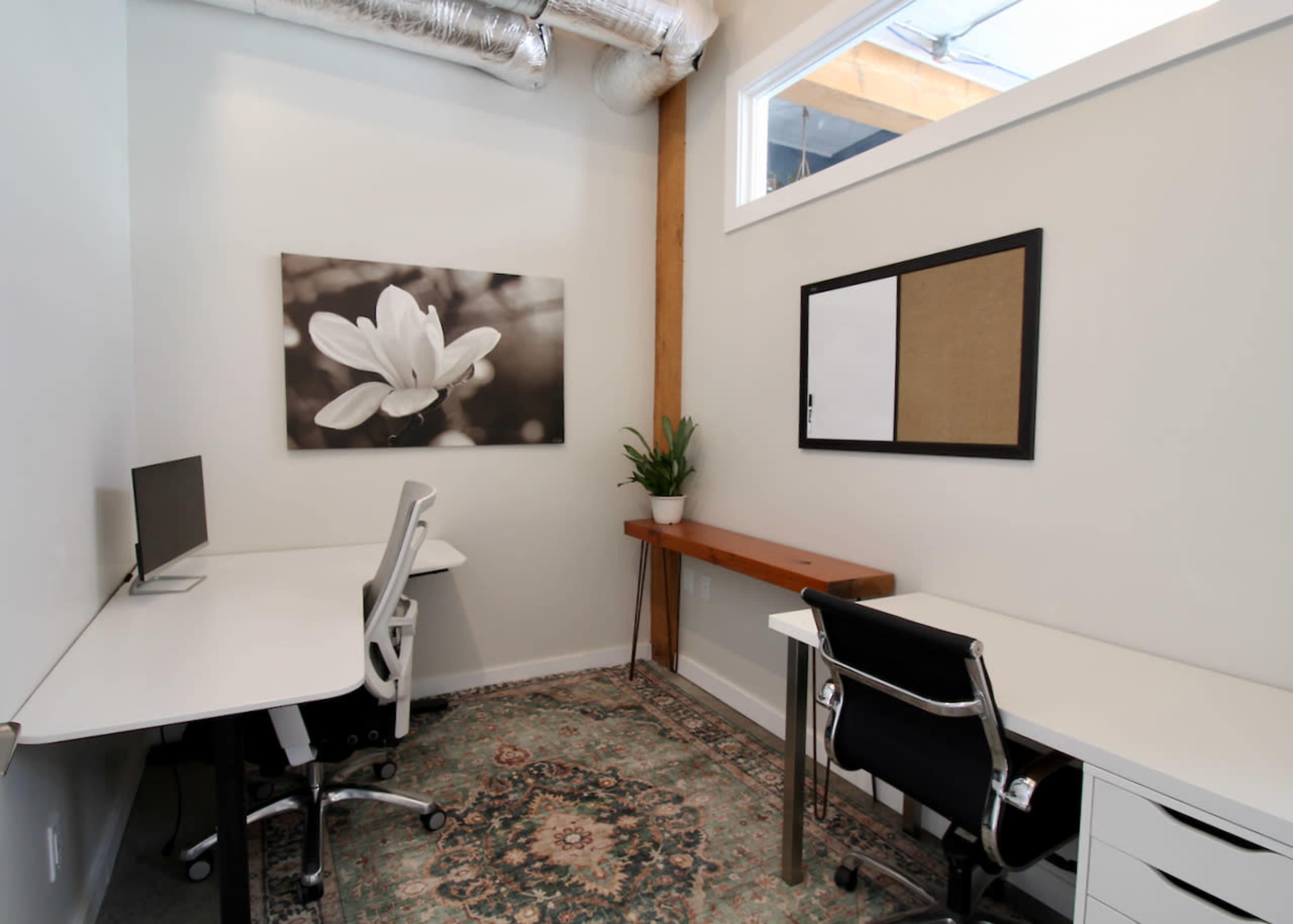 The image shows a small office space with two desks, a plant, and a wall-mounted picture of a flower.