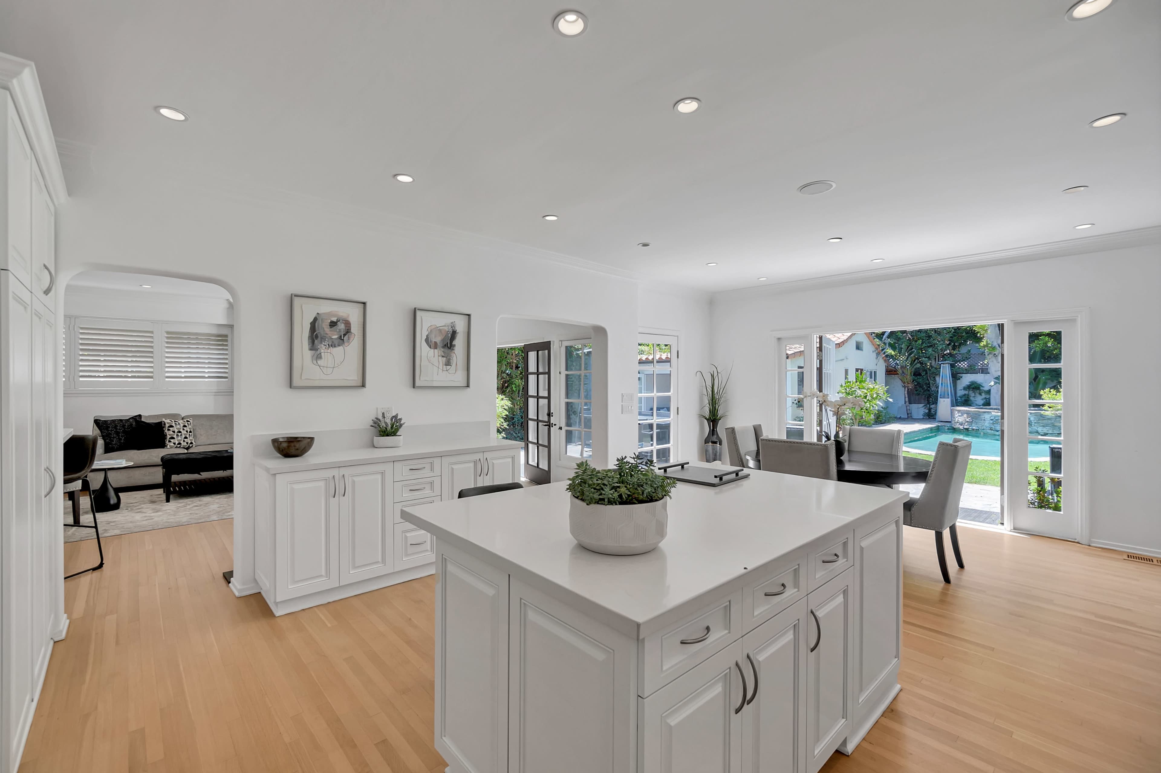 A bright kitchen with white cabinets and an island opens into a spacious living area featuring large windows overlooking a pool and outdoor seating area.