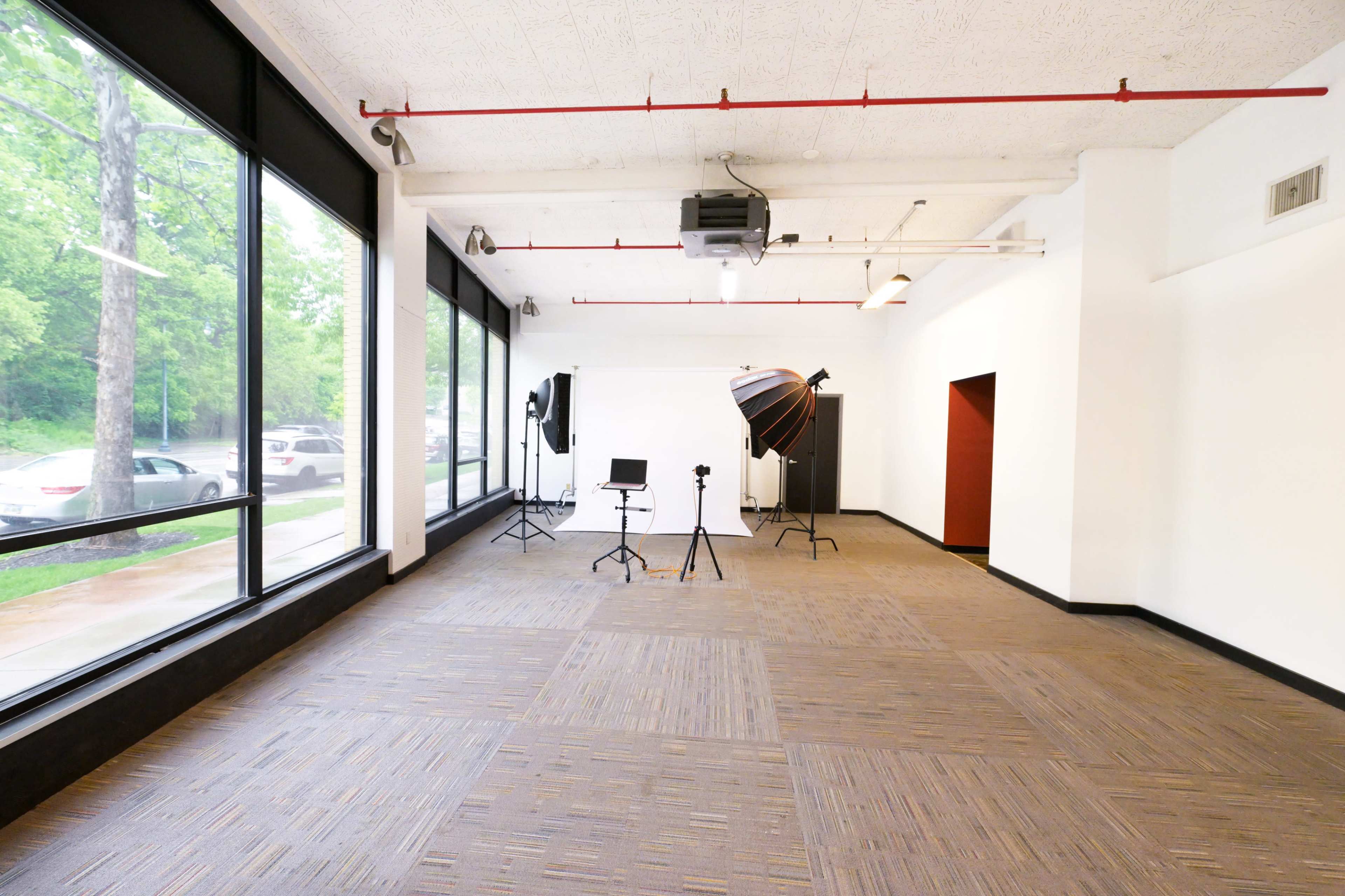 A spacious, empty studio features large windows, a gray carpet, and photography equipment set up in one corner.