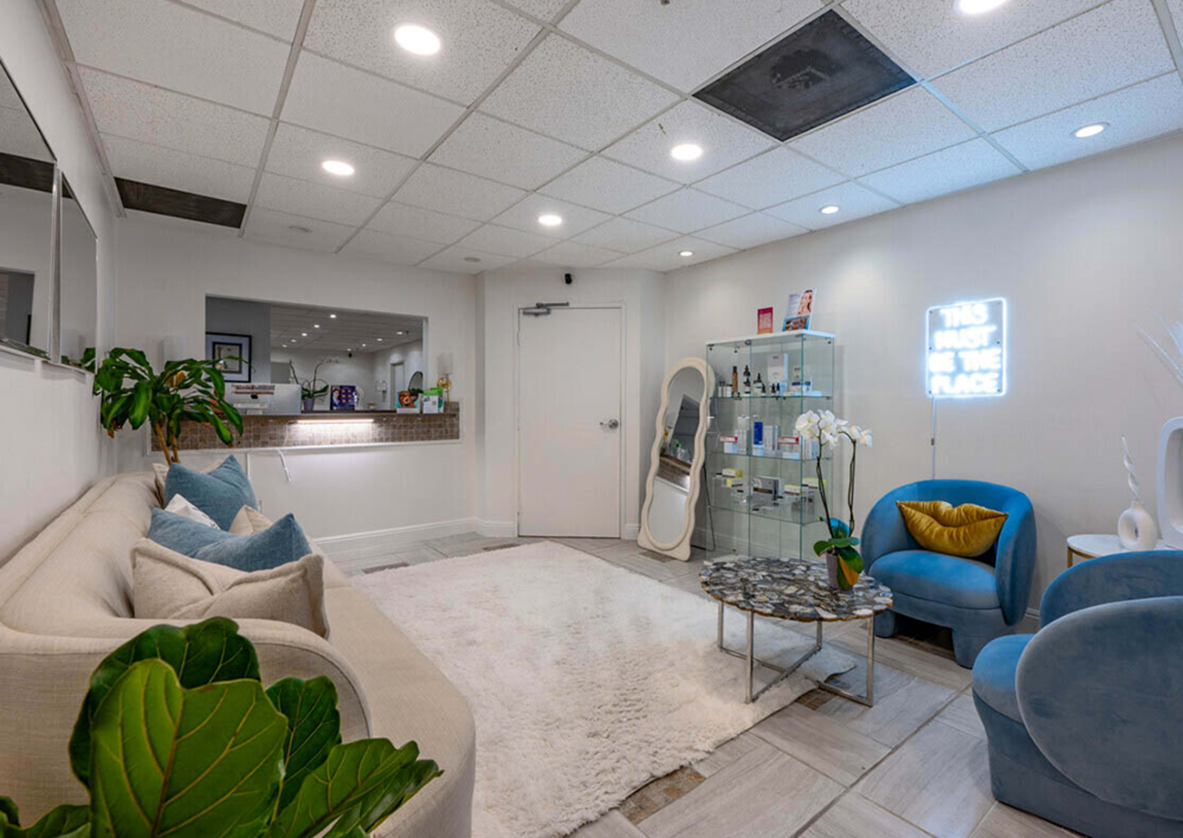 The image shows a modern waiting room with a large white rug, a sofa, blue accent chairs, and a neon sign on the wall.