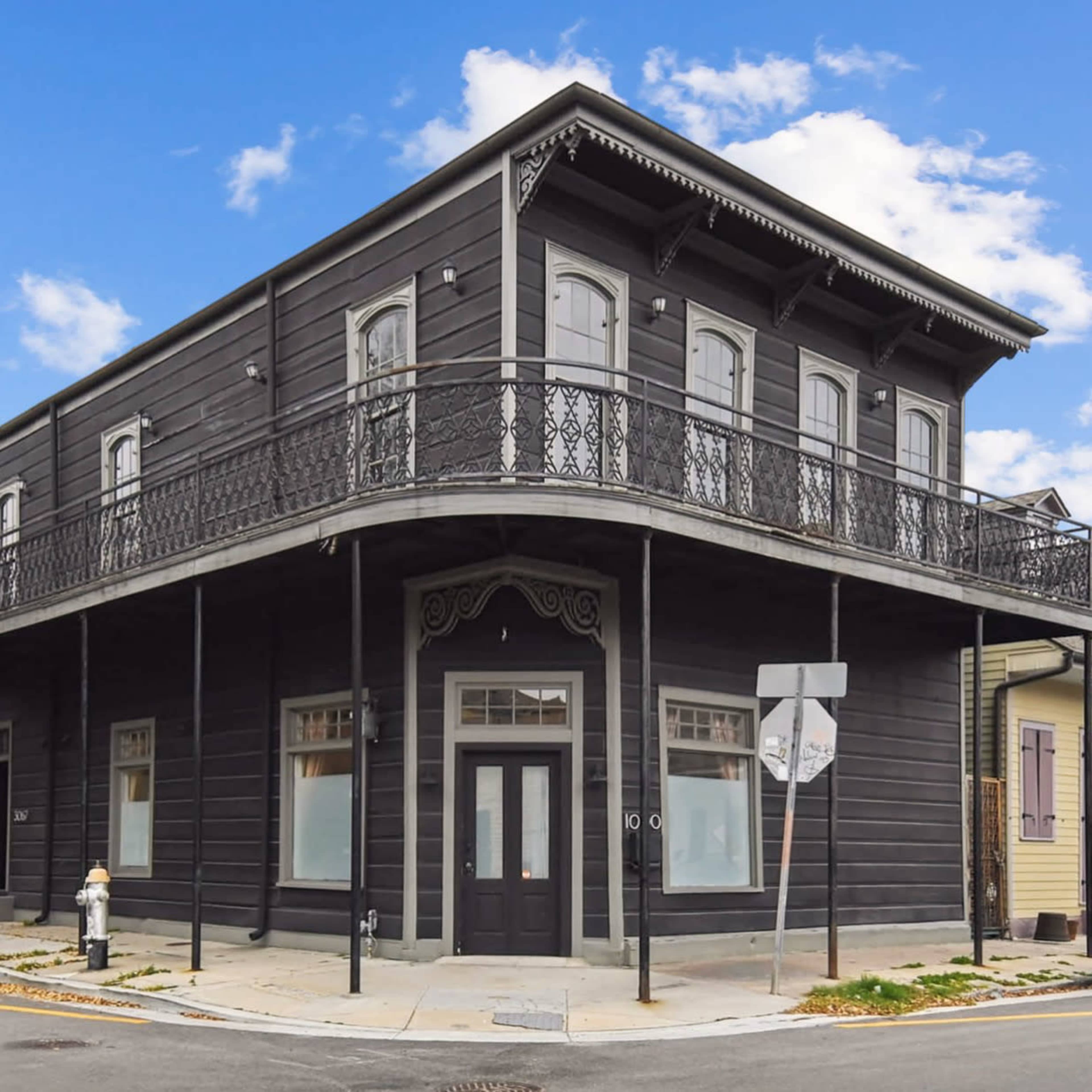 The image shows a two-story, dark-colored building with a decorative balcony and large windows located at a street corner.