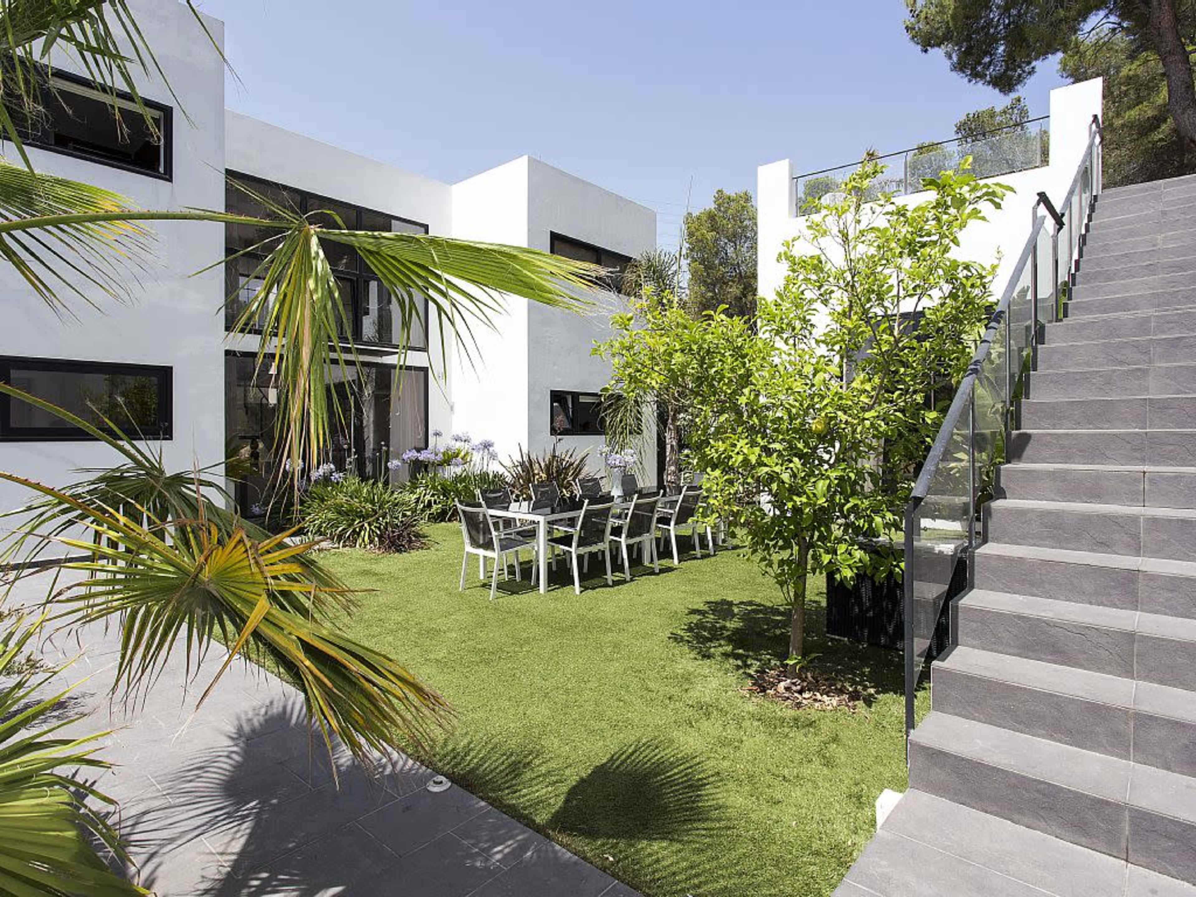 A modern home with large windows is surrounded by landscaped greenery and features a dining area with a table and chairs on the lawn, along with a staircase leading to an upper level.
