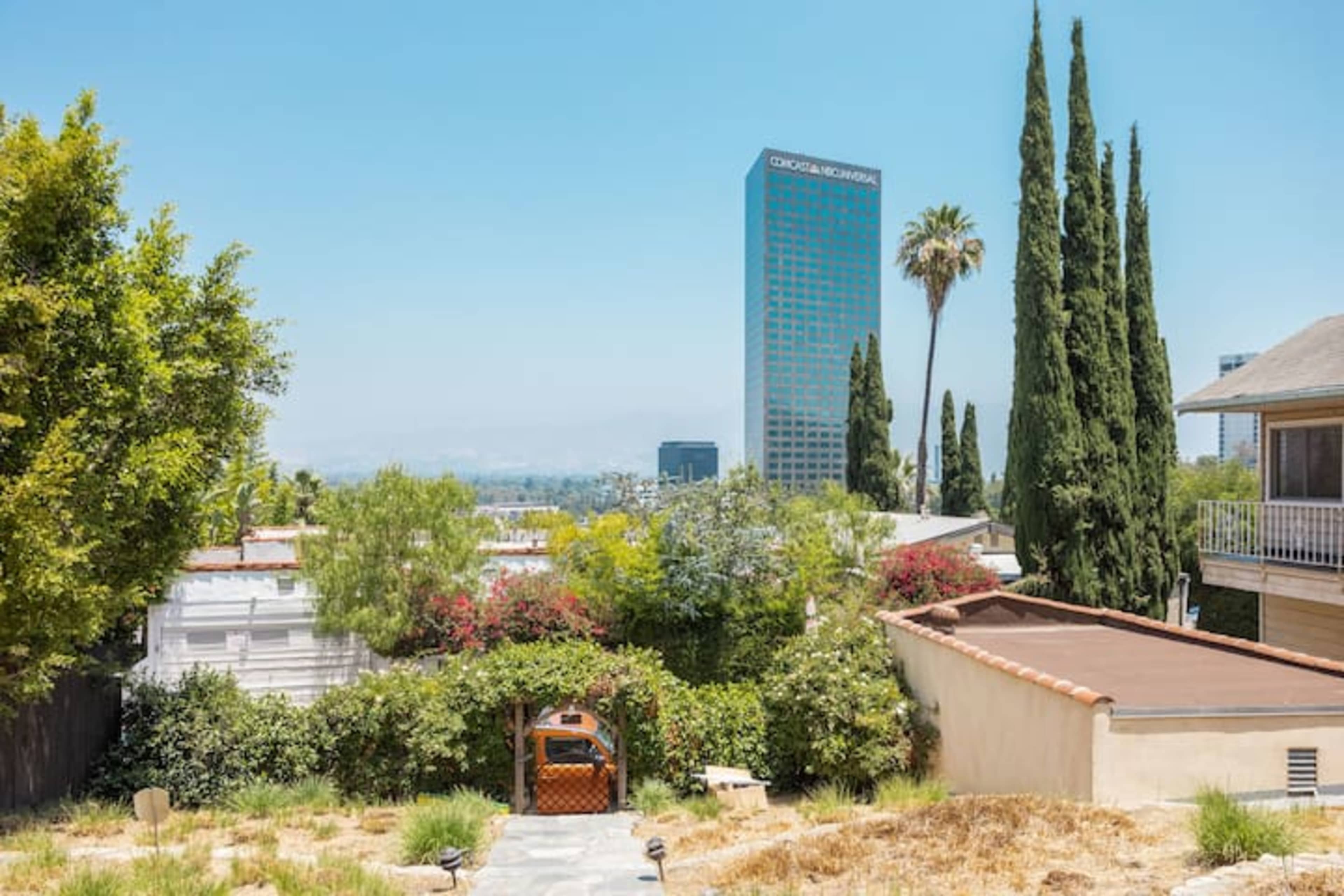 The image shows a suburban landscape with a stone pathway leading to a wooden gazebo, surrounded by greenery, with tall buildings visible in the background.