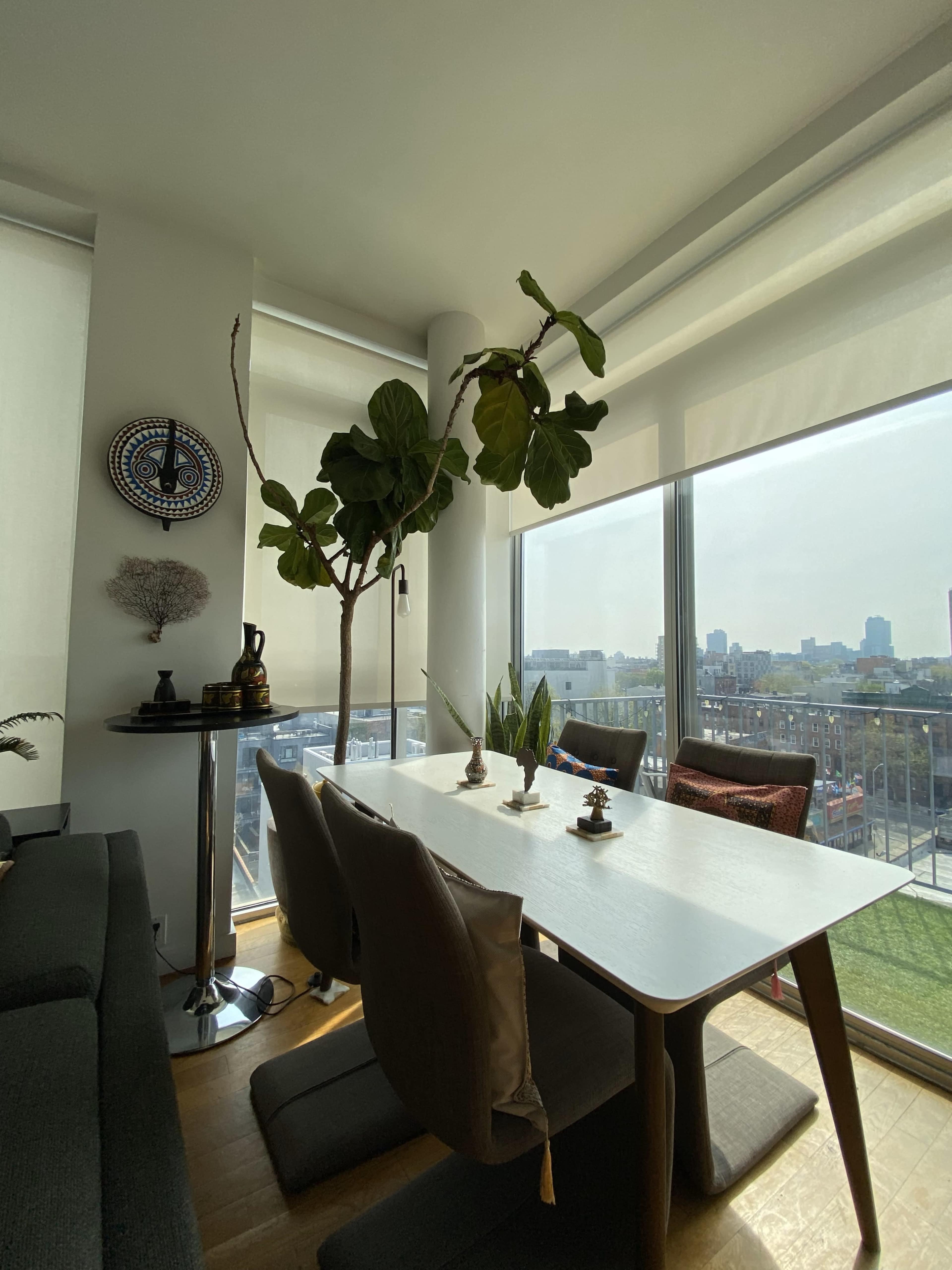 The image depicts a modern dining area with a table set for four, large windows providing natural light, and a potted tree in the corner.