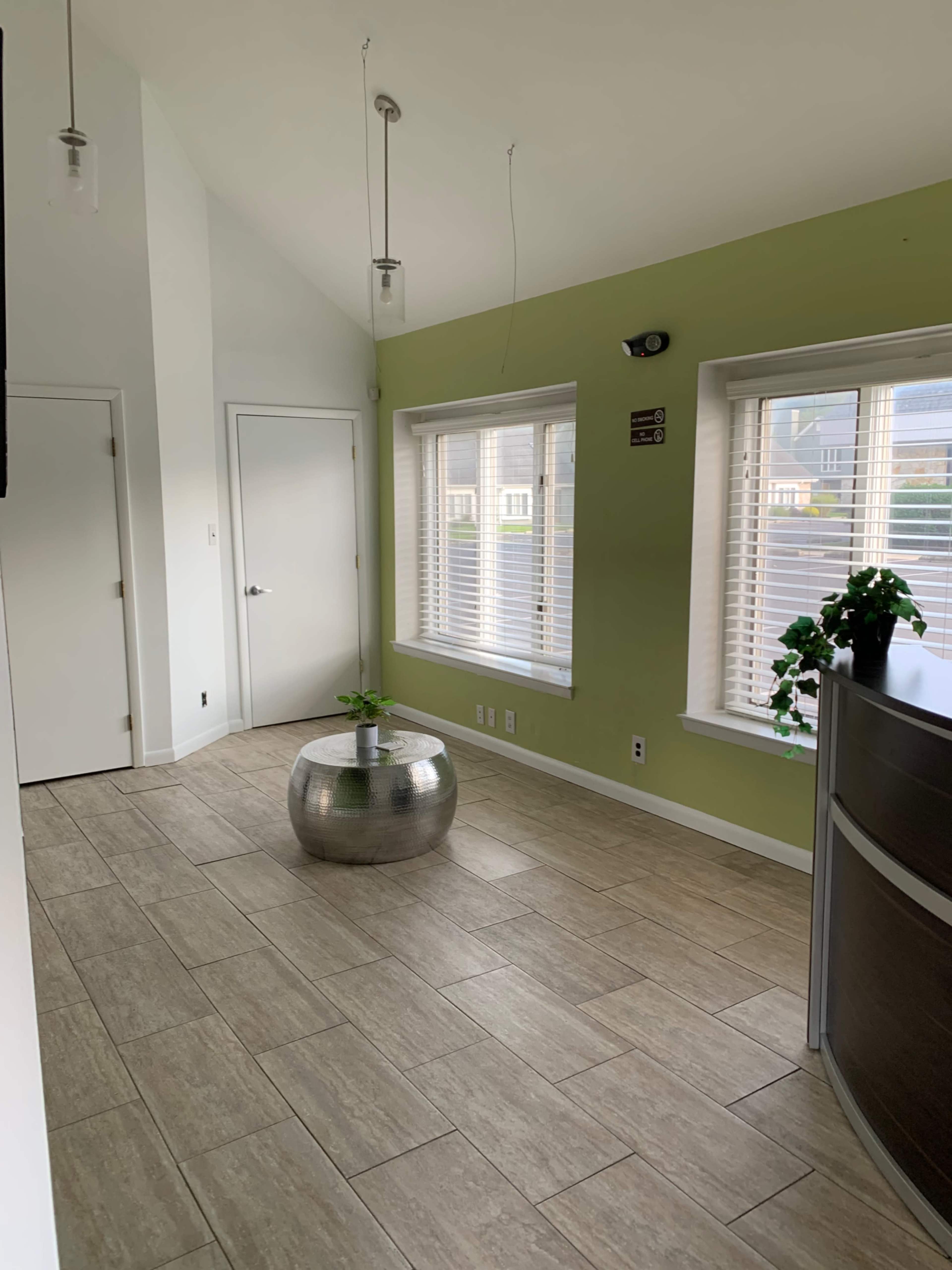 A modern, well-lit interior features a green accent wall, large windows with blinds, and a round metallic table with a small plant.