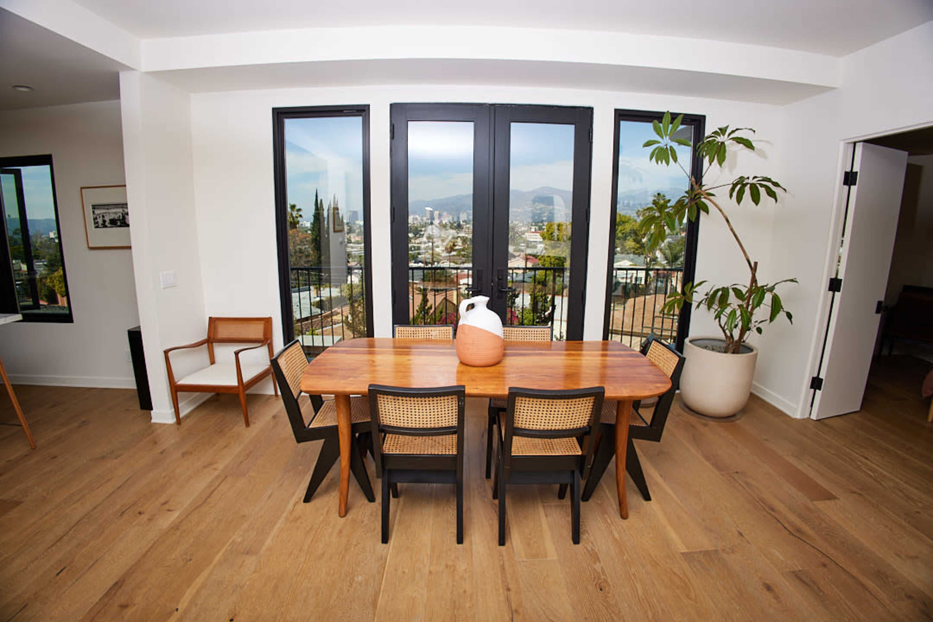 A wooden dining table with black chairs is set in a well-lit room featuring large windows that overlook a cityscape and mountains.