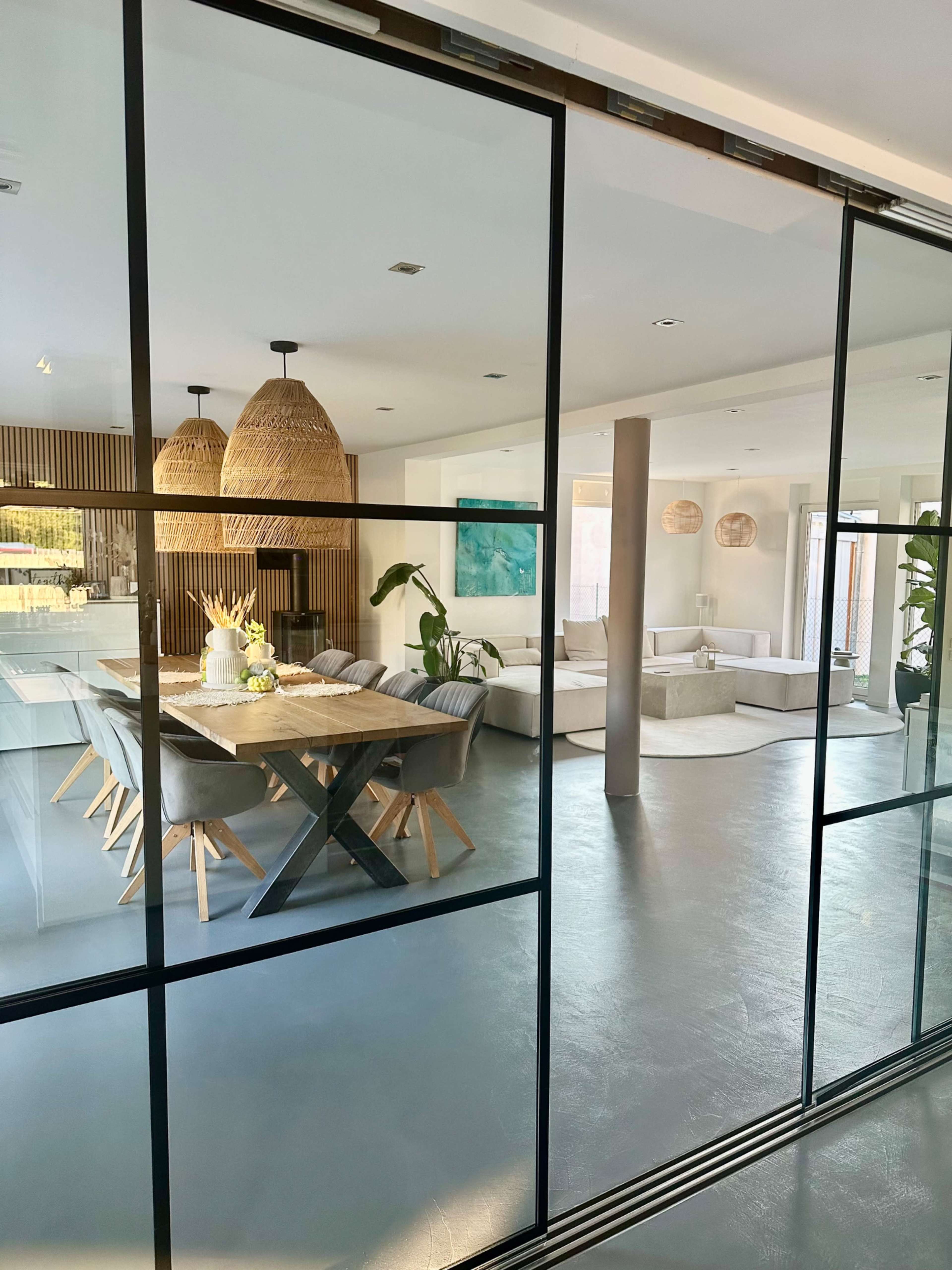 A modern open-concept dining area features a large wooden table, comfortable seating, and pendant lighting, with a view from an adjacent room through a glass partition.