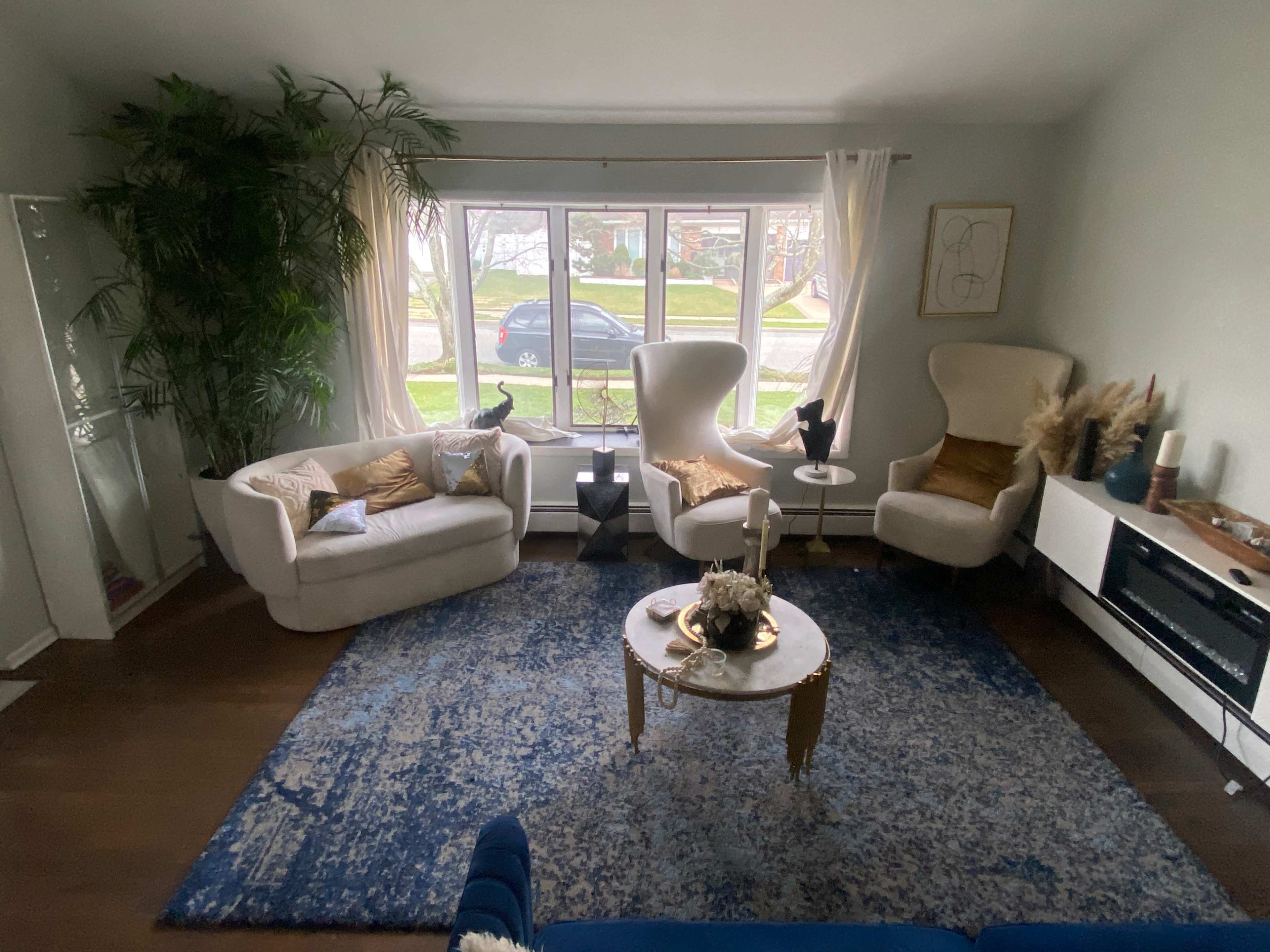 A cozy living room features two armchairs, a white sofa, a round coffee table, and a large window with a view of the street outside.