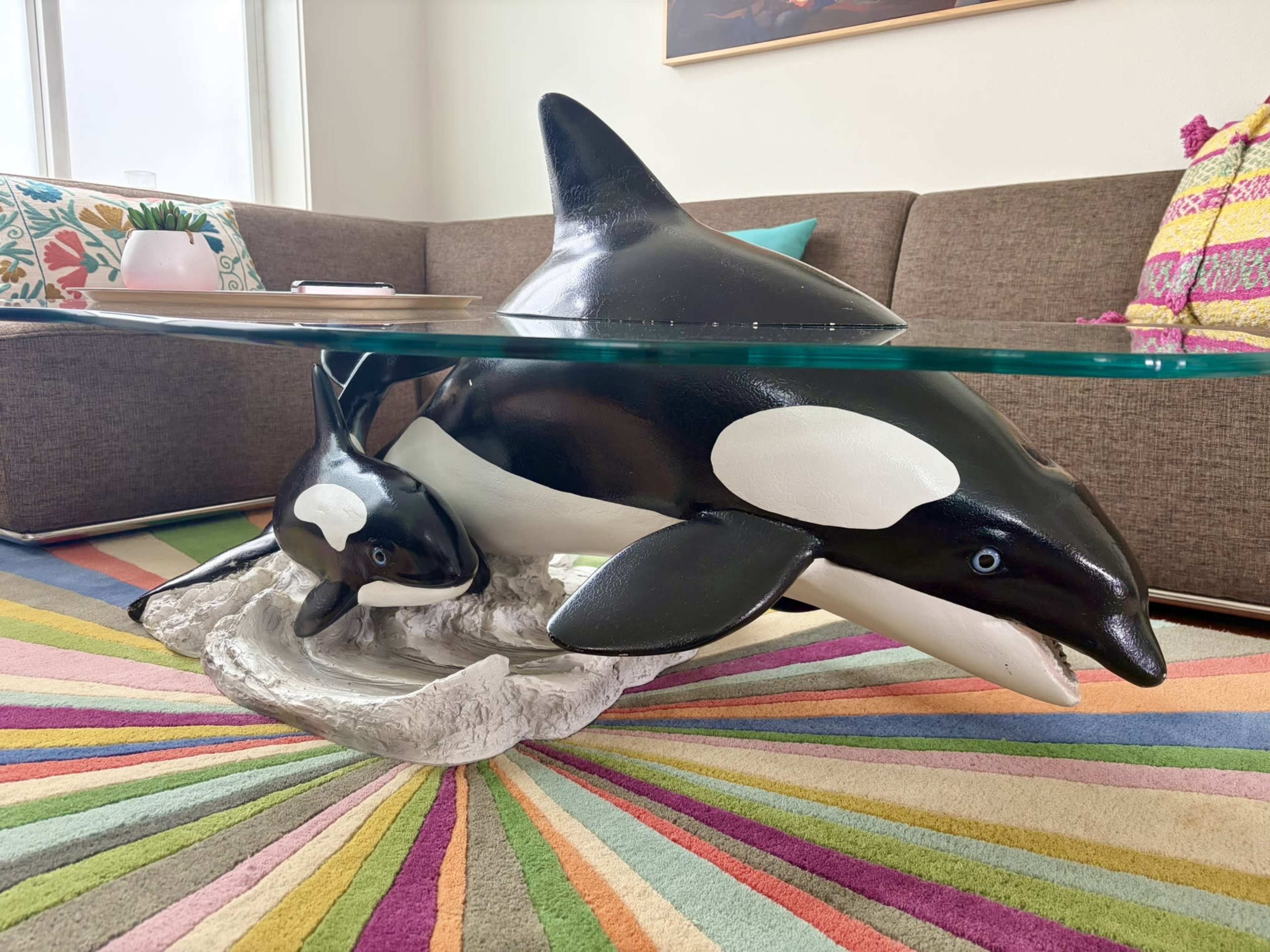 The image shows a coffee table featuring a sculpture of two orcas beneath a glass top, positioned on a colorful, striped rug in a living room setting.