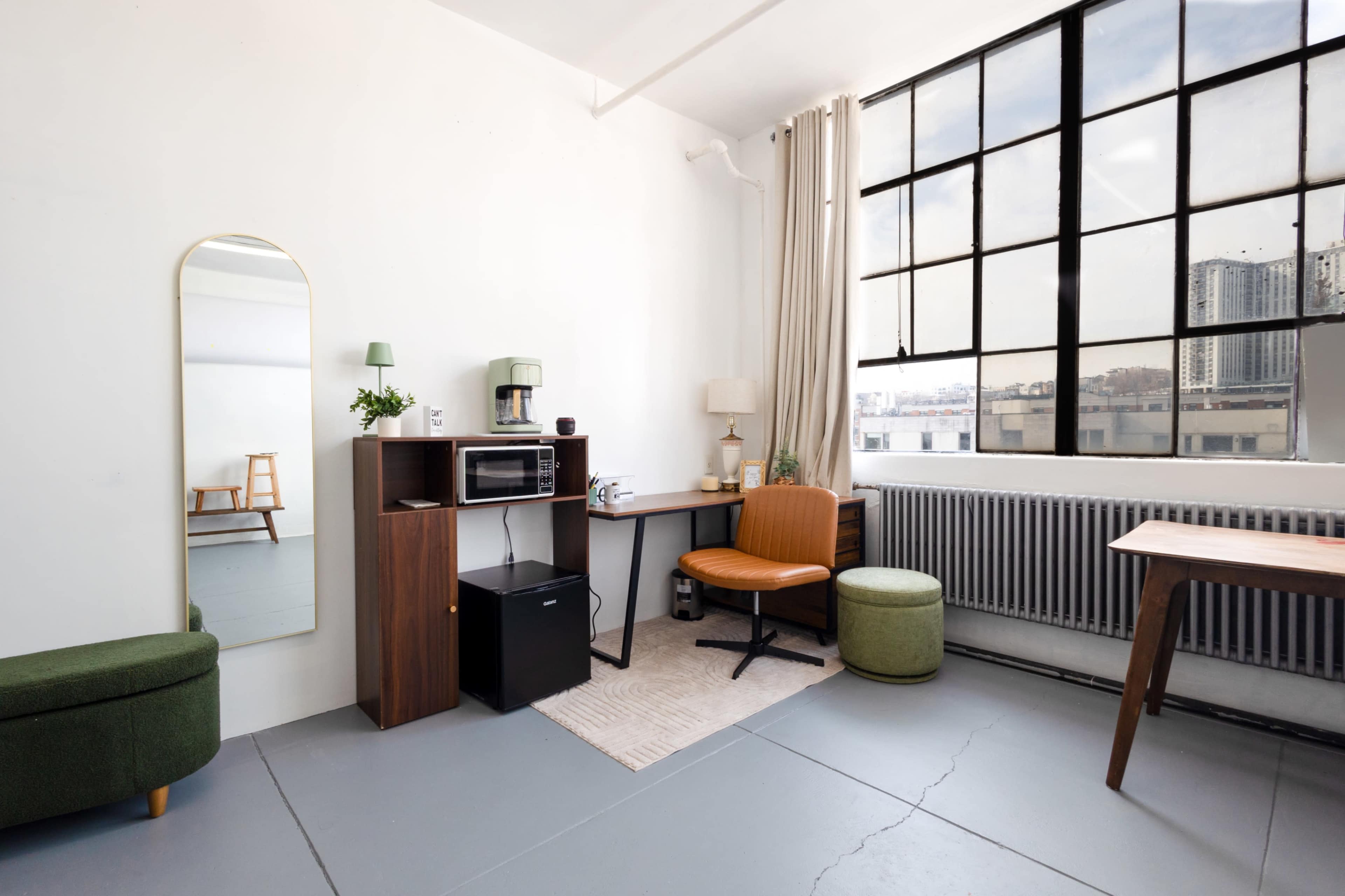 The image shows a minimalist studio space featuring a desk, a seating area, a mirror, and a large window with city views.