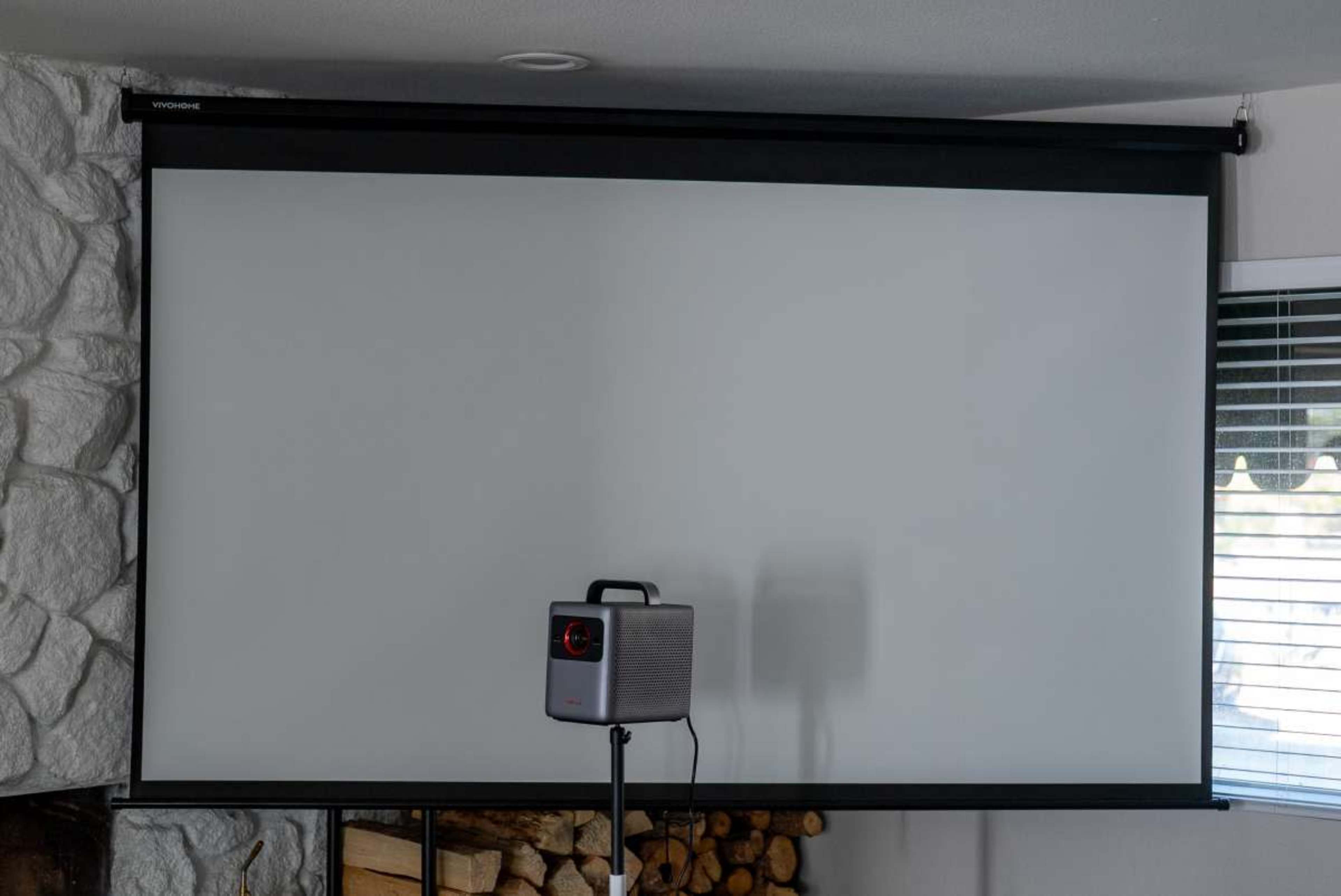 The image shows a wall-mounted projector screen in a room, accompanied by a projector on a stand in front of it.