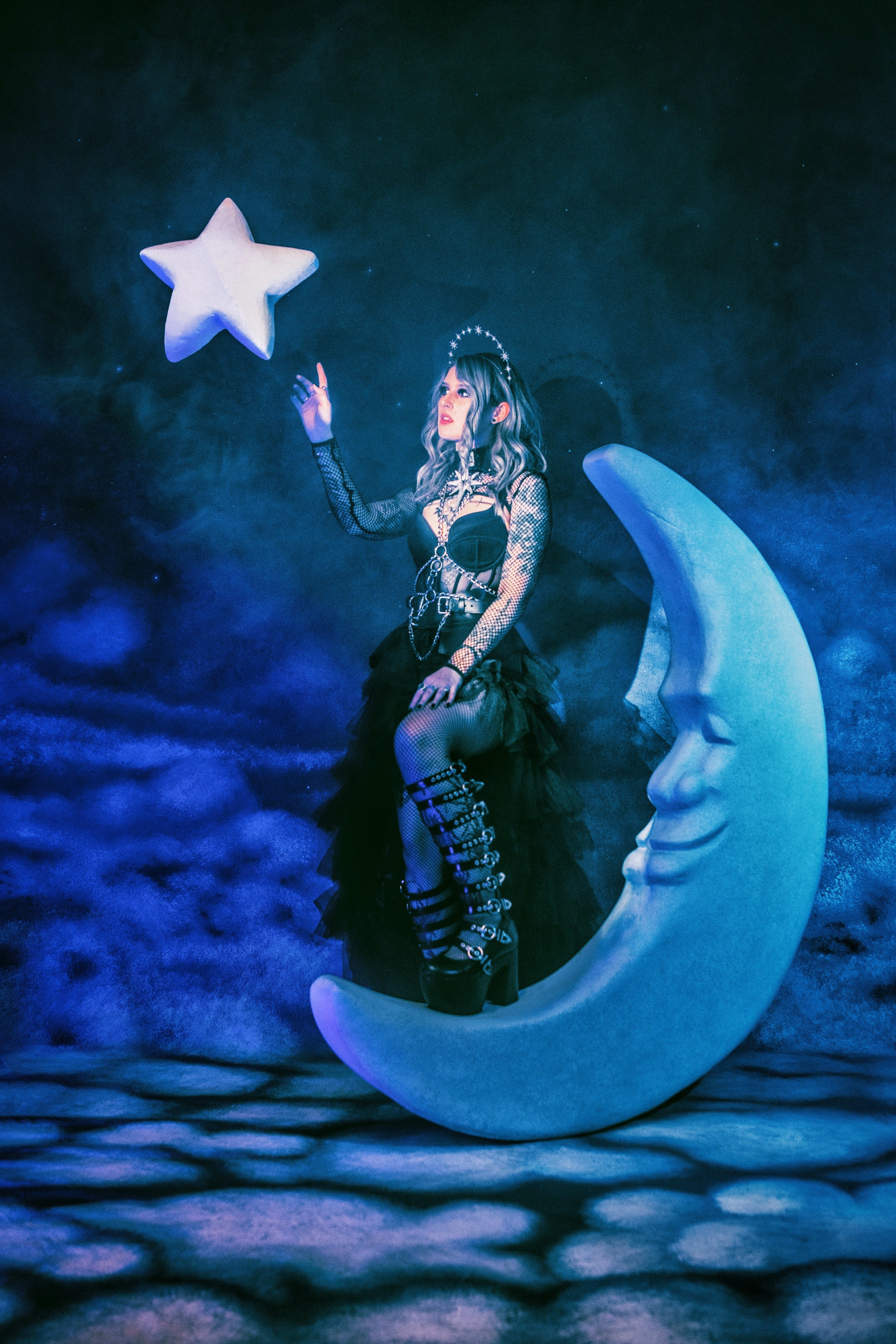 A fashion photoshoot featuring a woman reaching to a star while sitting on the blue-lit moon.