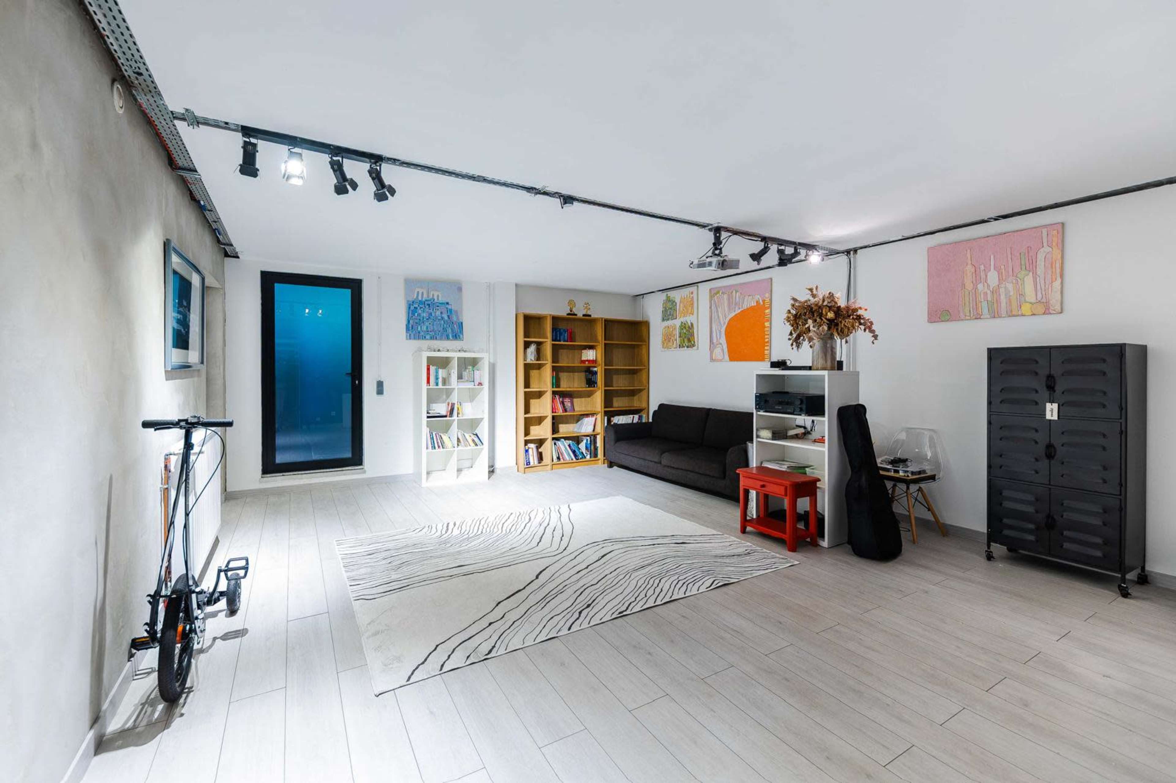 The image shows a modern interior space with a gray wall, light wooden flooring, a black sofa, a bookshelf, and various pieces of artwork on the walls.