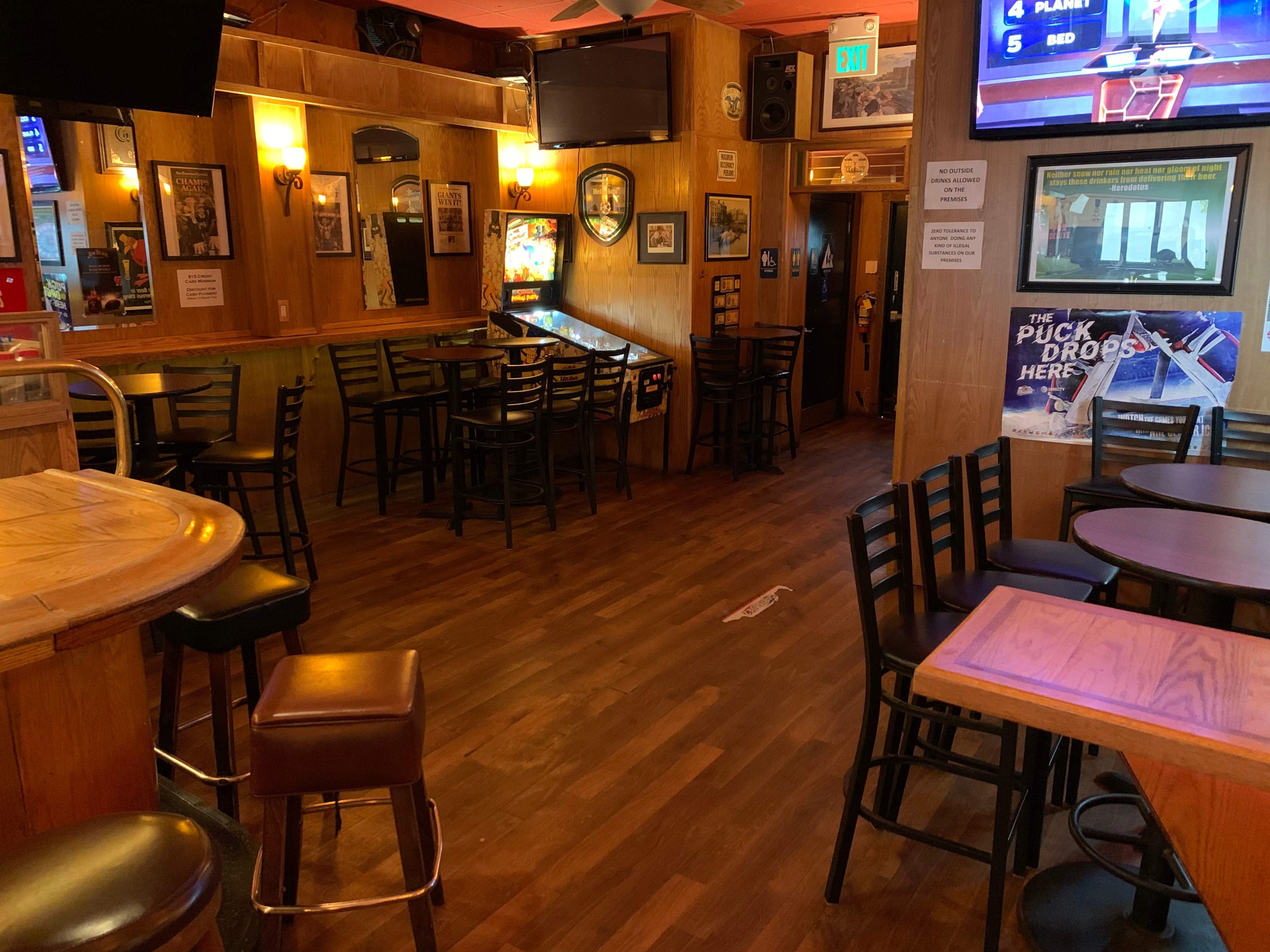 The image shows a dimly lit bar area with wooden walls, empty tables, and a pinball machine in the background.
