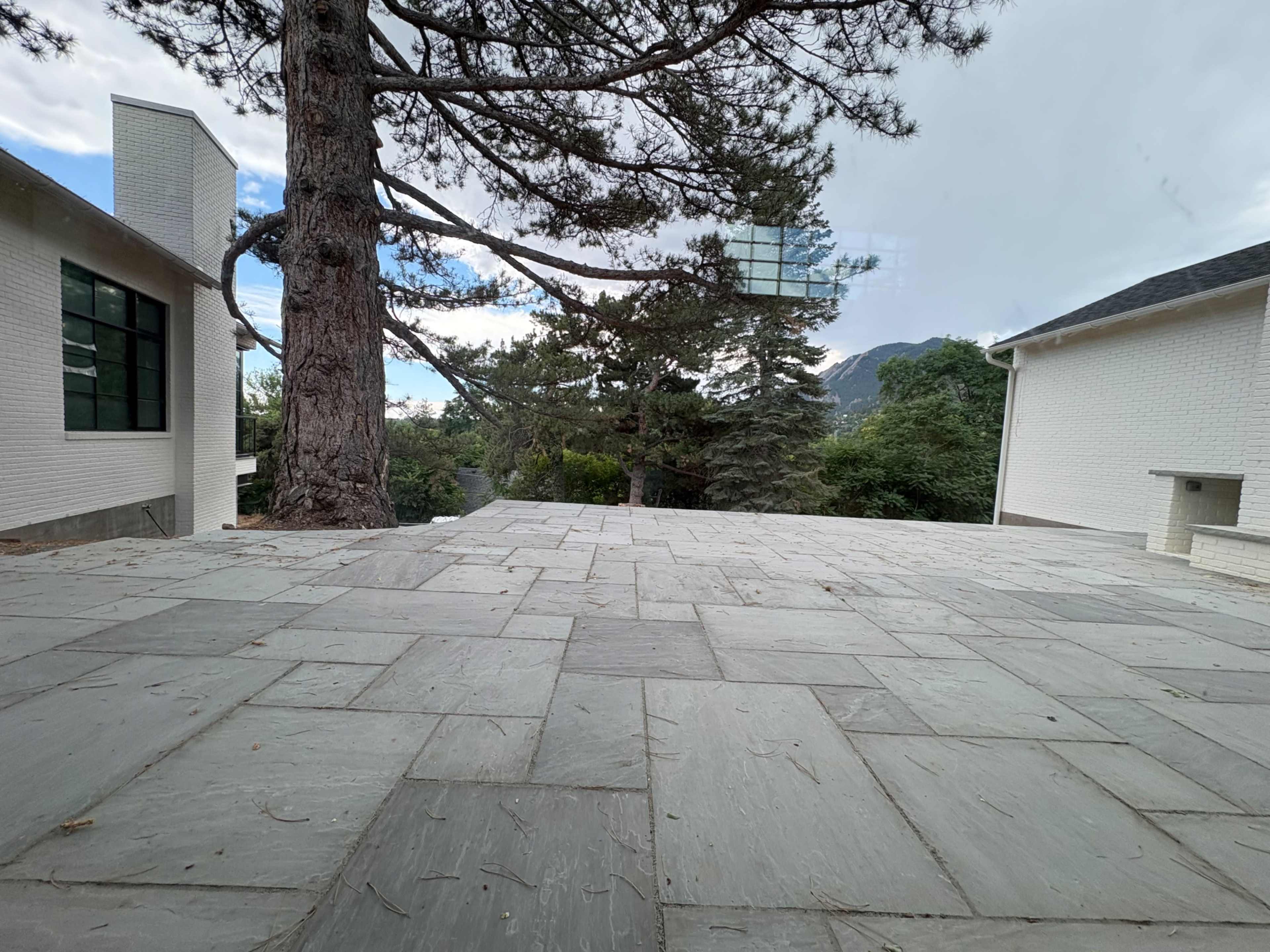The image shows a spacious stone patio surrounded by trees and two white buildings.
