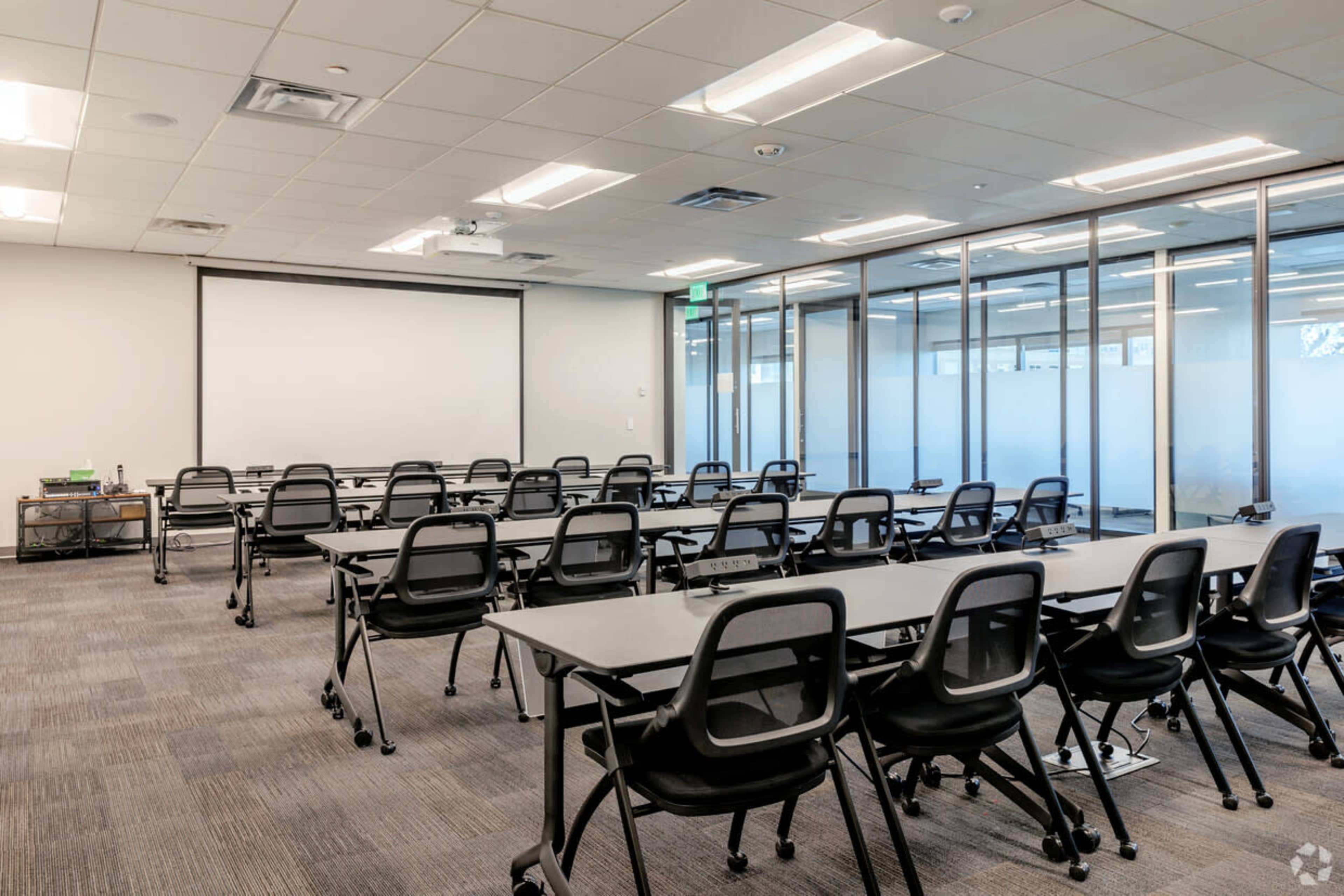 A modern meeting room features rows of tables and chairs arranged for a presentation, with a large screen at the front and glass walls on one side.