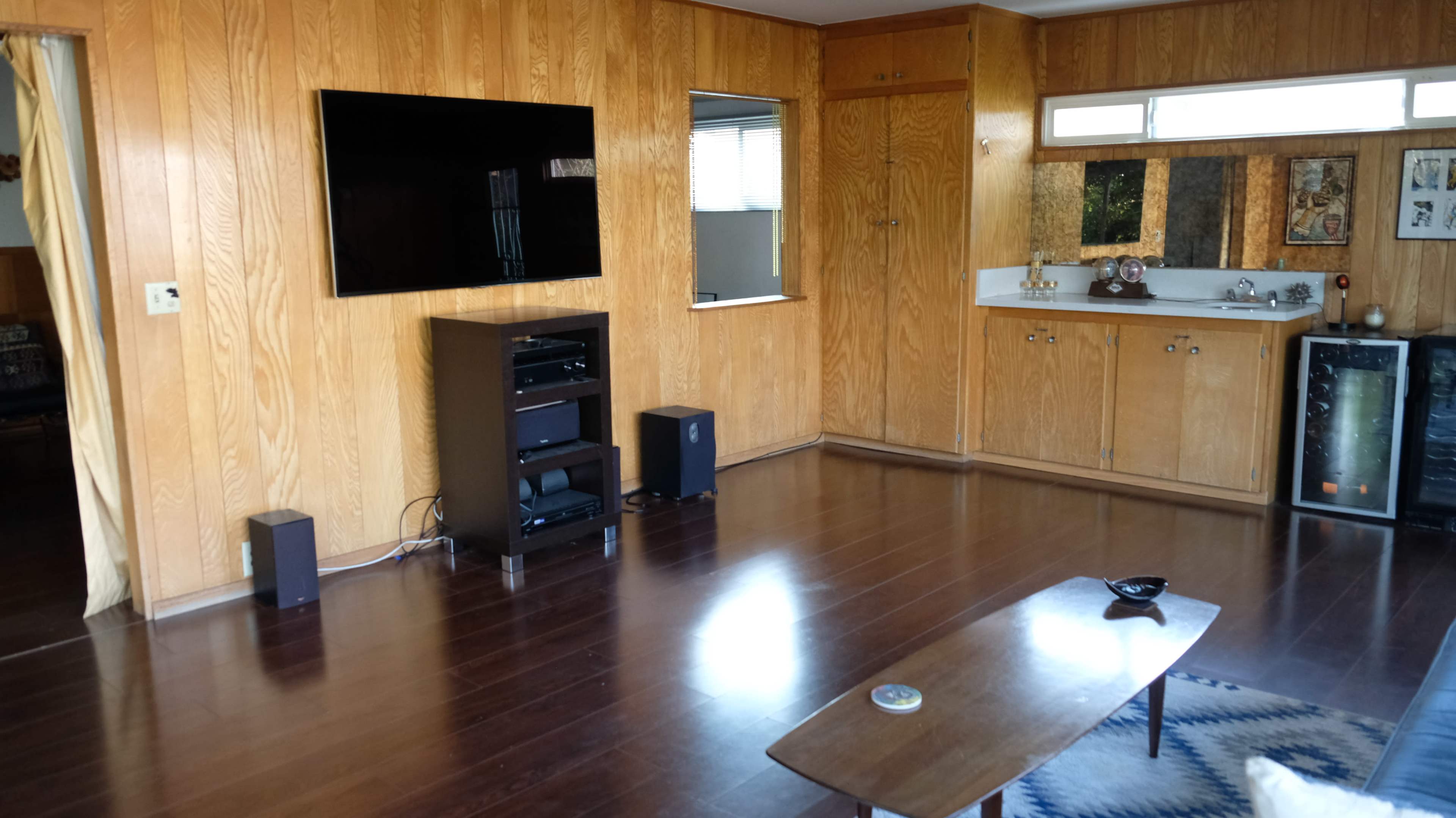The image shows a living room with wooden paneling, a wall-mounted TV, a black entertainment unit, and a small coffee table on a patterned rug.