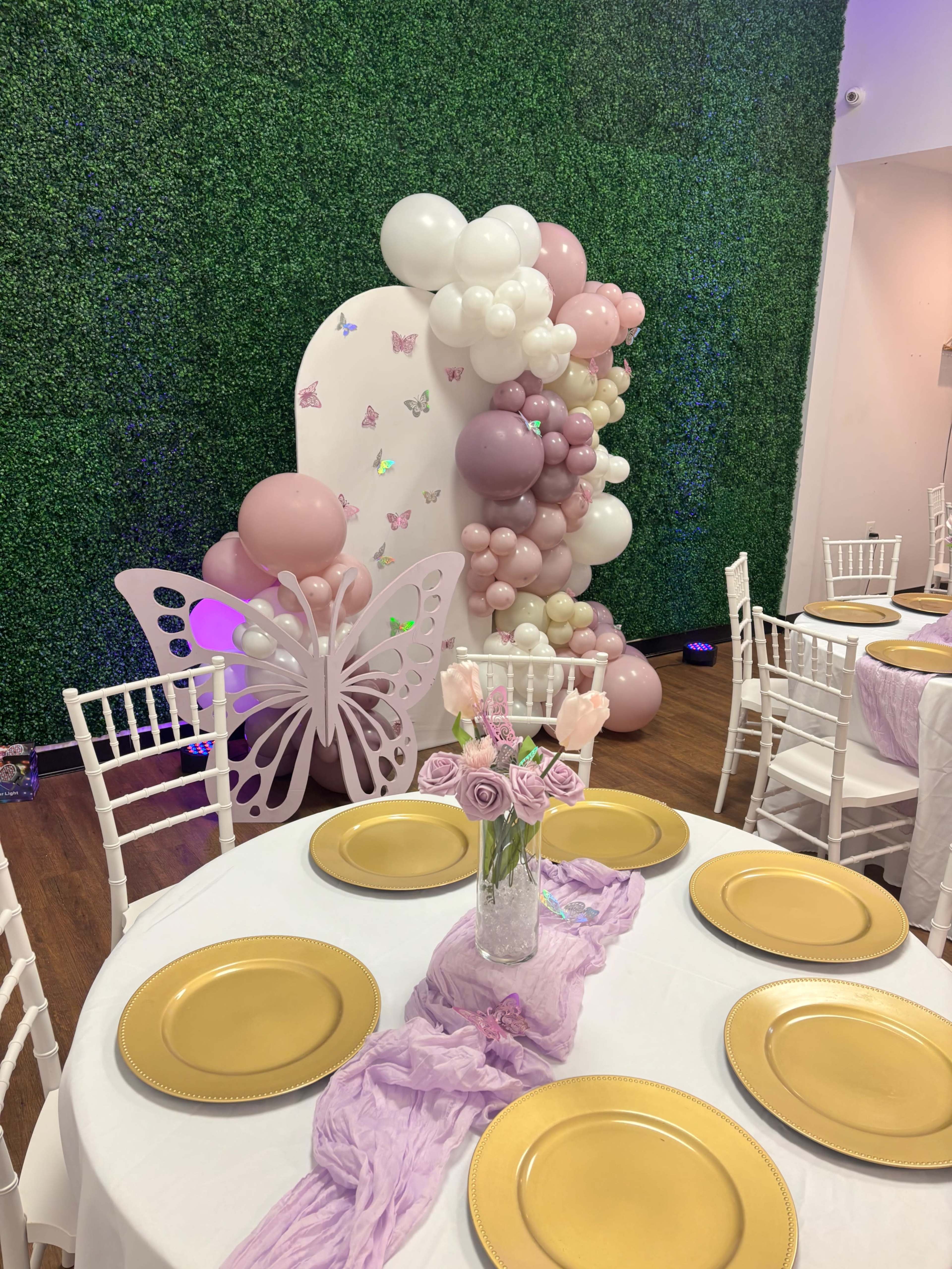 A decorated event space features white tables with gold plates, a floral centerpiece, and a backdrop of greenery adorned with balloons in shades of pink and purple.