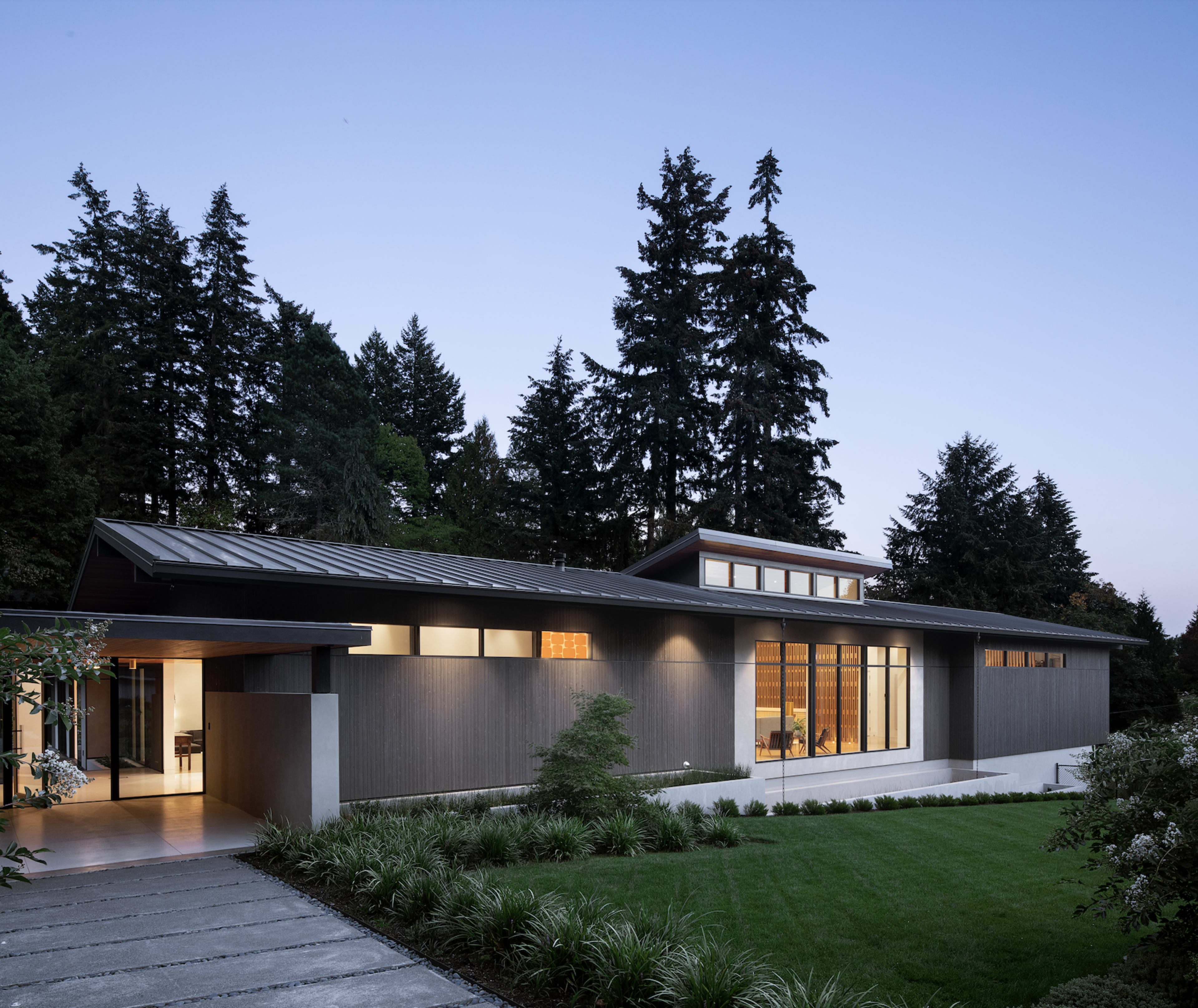 A modern house with a flat roof and large windows is situated amidst tall trees, featuring a landscaped front yard with greenery.