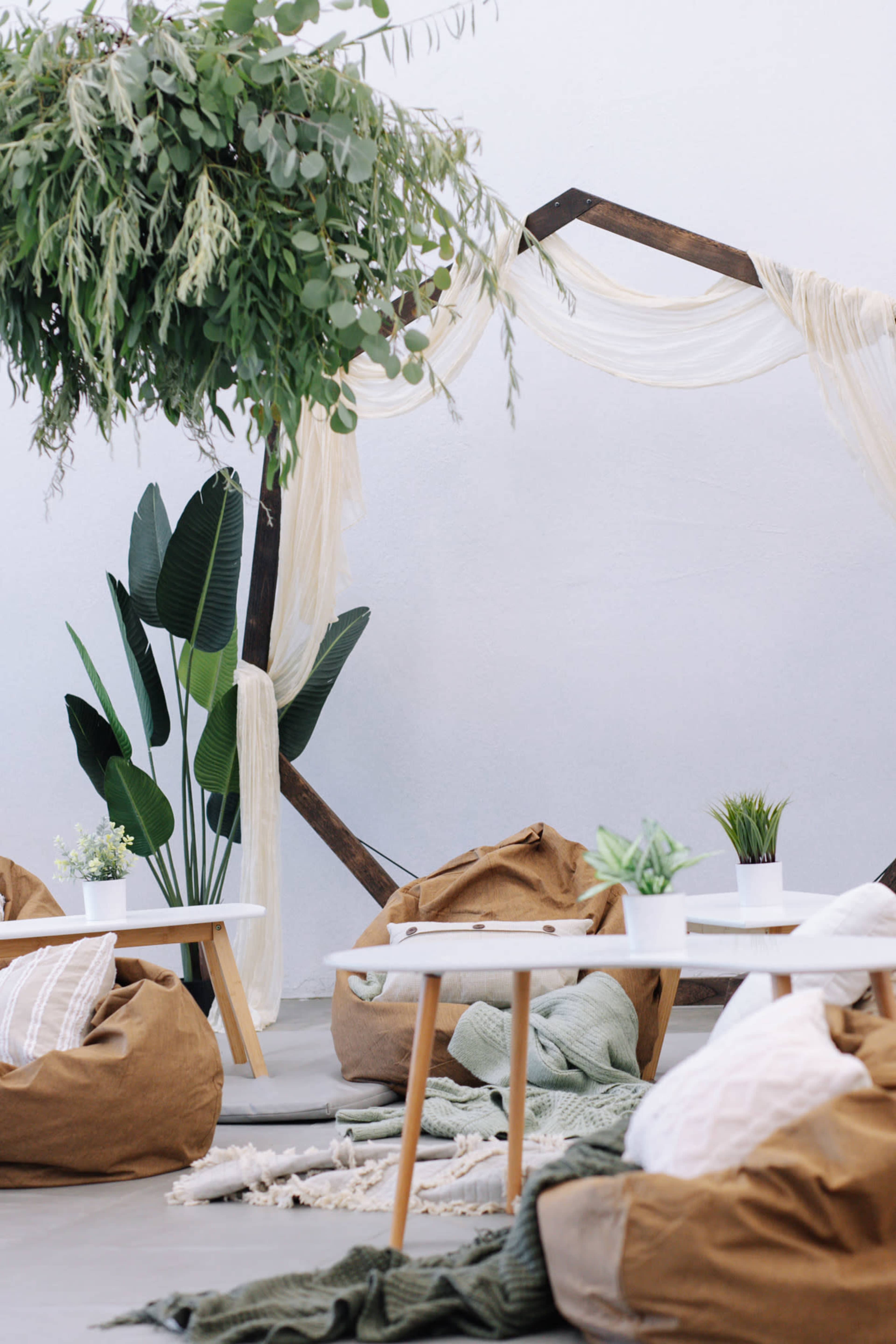 A cozy indoor space features bean bags, potted plants, and a decorative wooden structure draped with fabric and greenery.