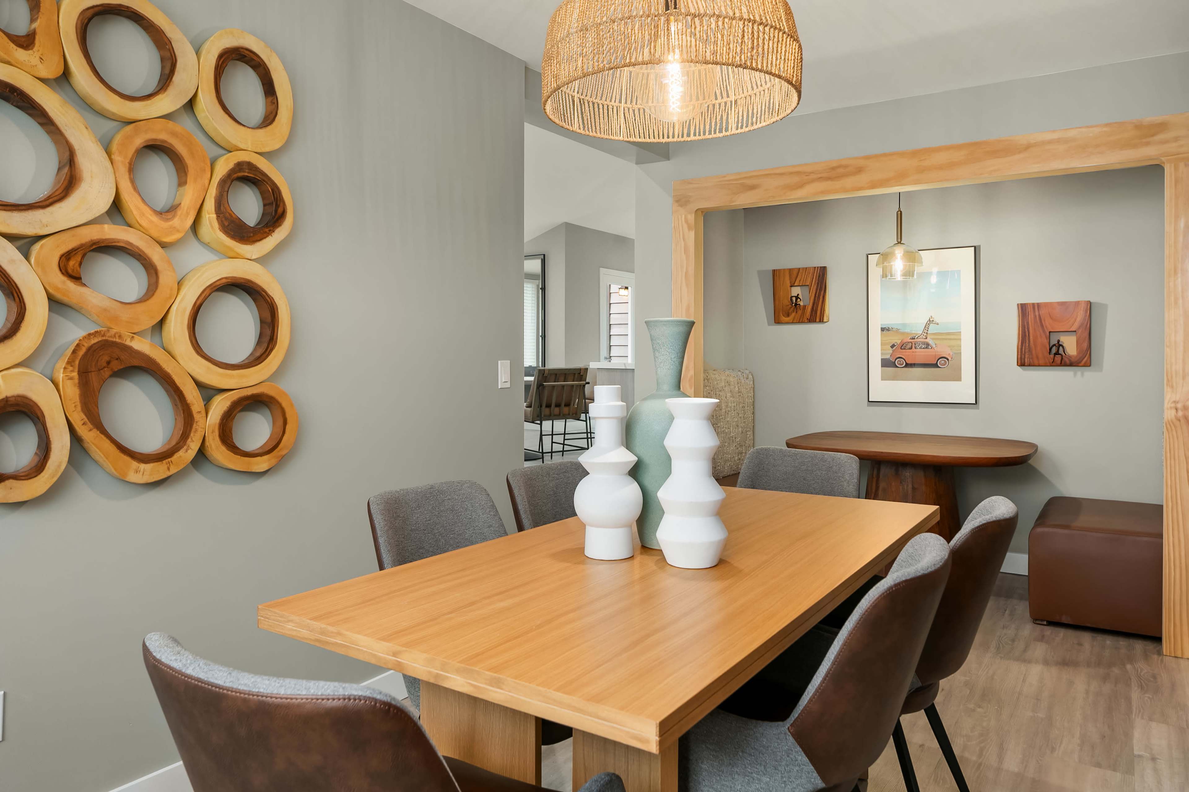 The image shows a modern dining area featuring a wooden table surrounded by upholstered chairs, a decorative wall with circular wood accents, and various art pieces.