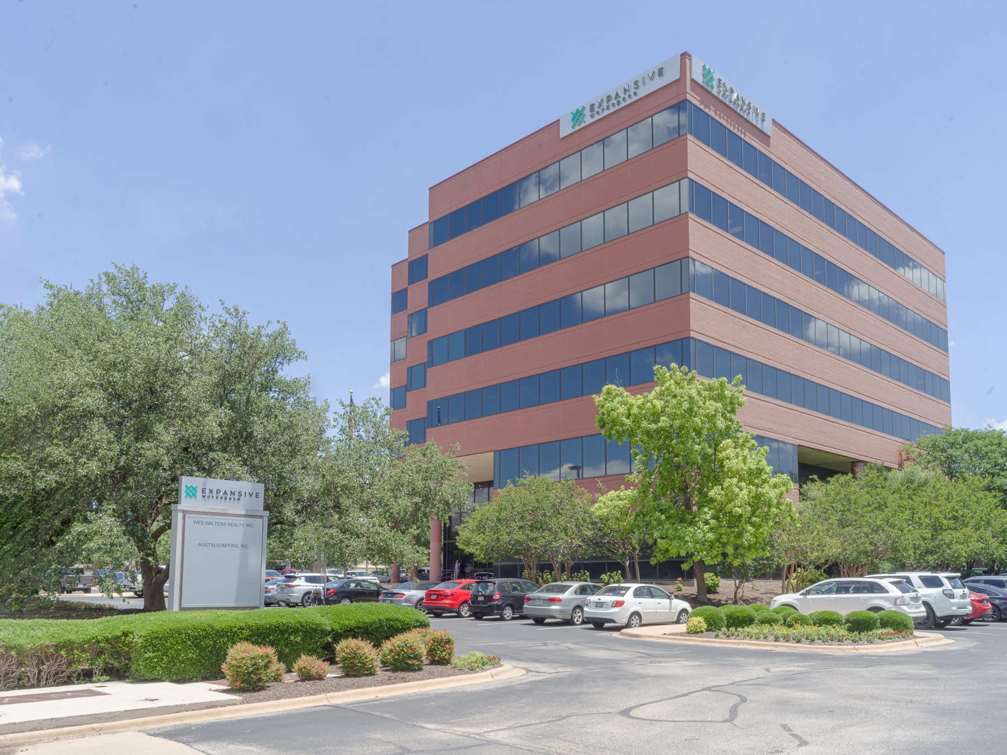 A modern multi-story office building with large windows and landscaping surrounds it, located in a parking lot area.