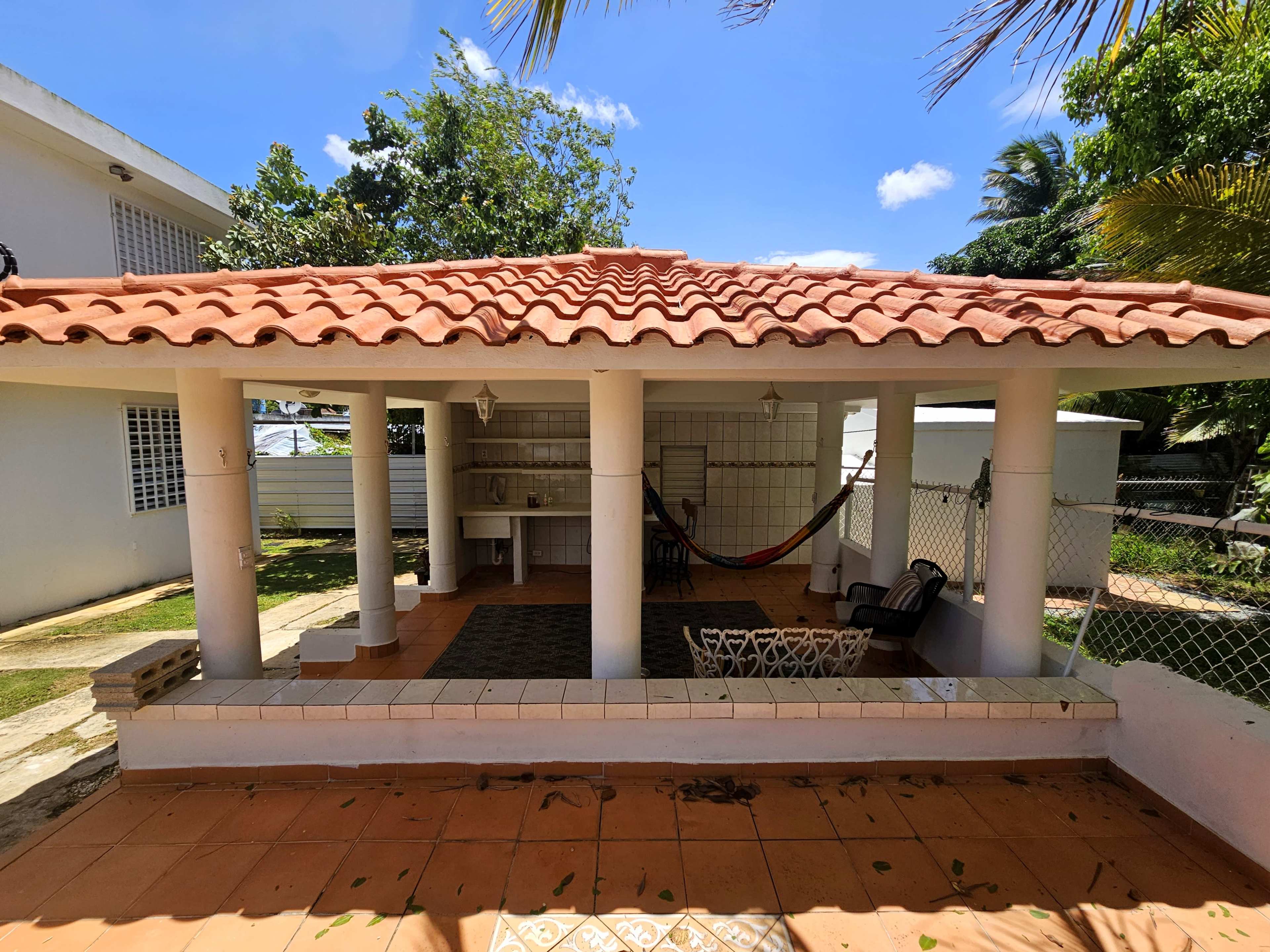 The image shows a patio area featuring a tiled floor, columns, and a roof with red tiles, surrounded by greenery and a hammock.