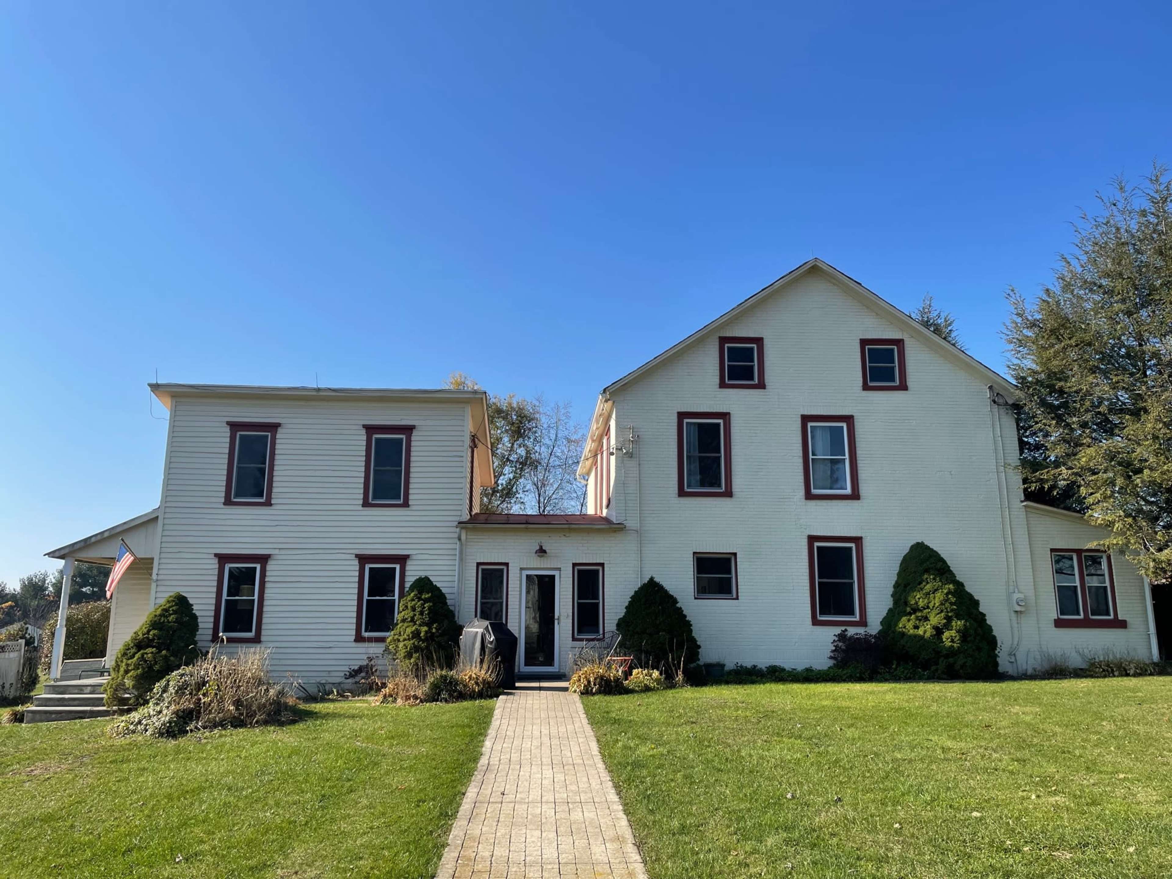 The image shows a two-story house with a combination of white siding and a red-trimmed facade, featuring a symmetrical layout and a paved pathway leading to the entrance.
