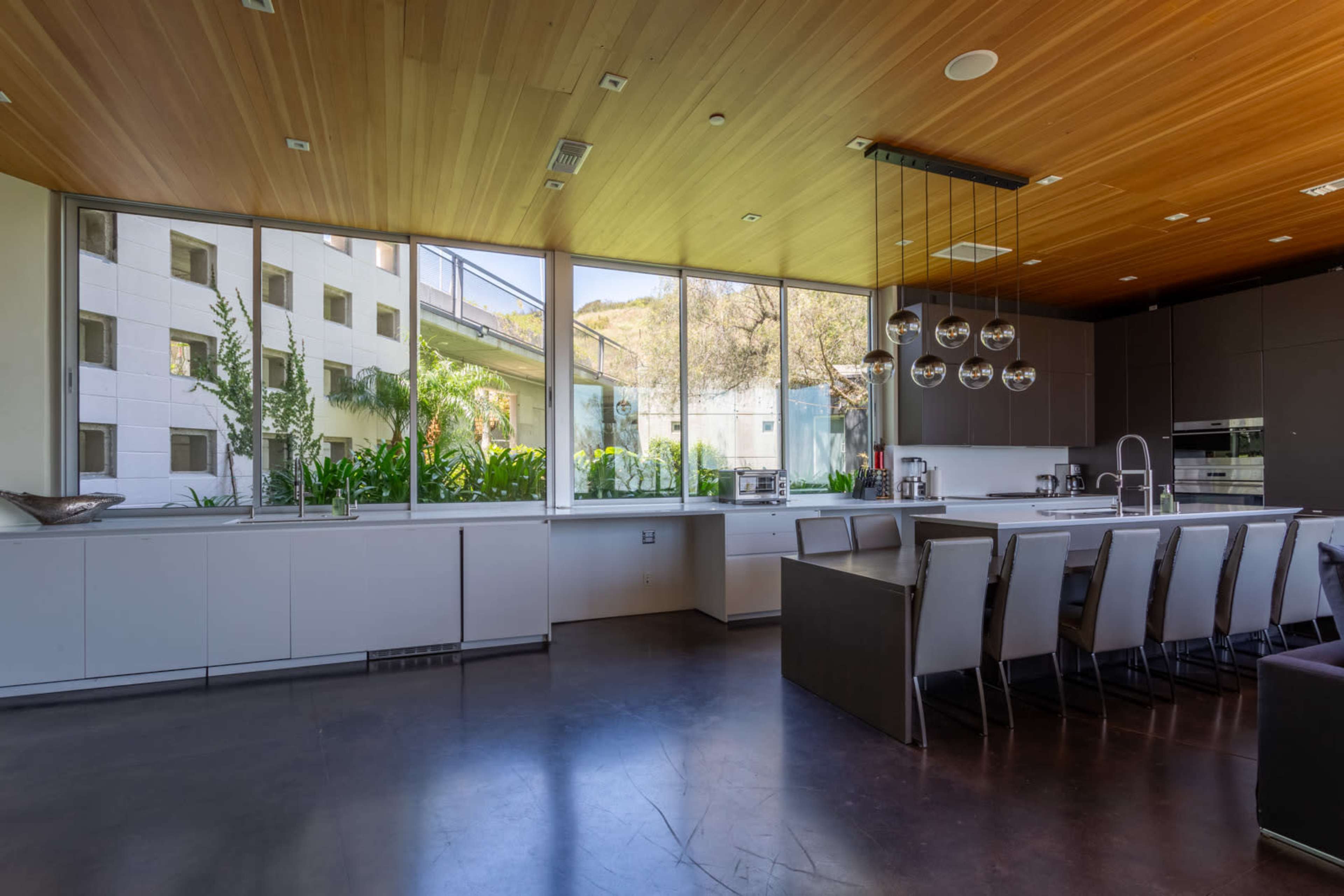 A modern kitchen features large windows with views of greenery, a central island, and a dining table with eight chairs.