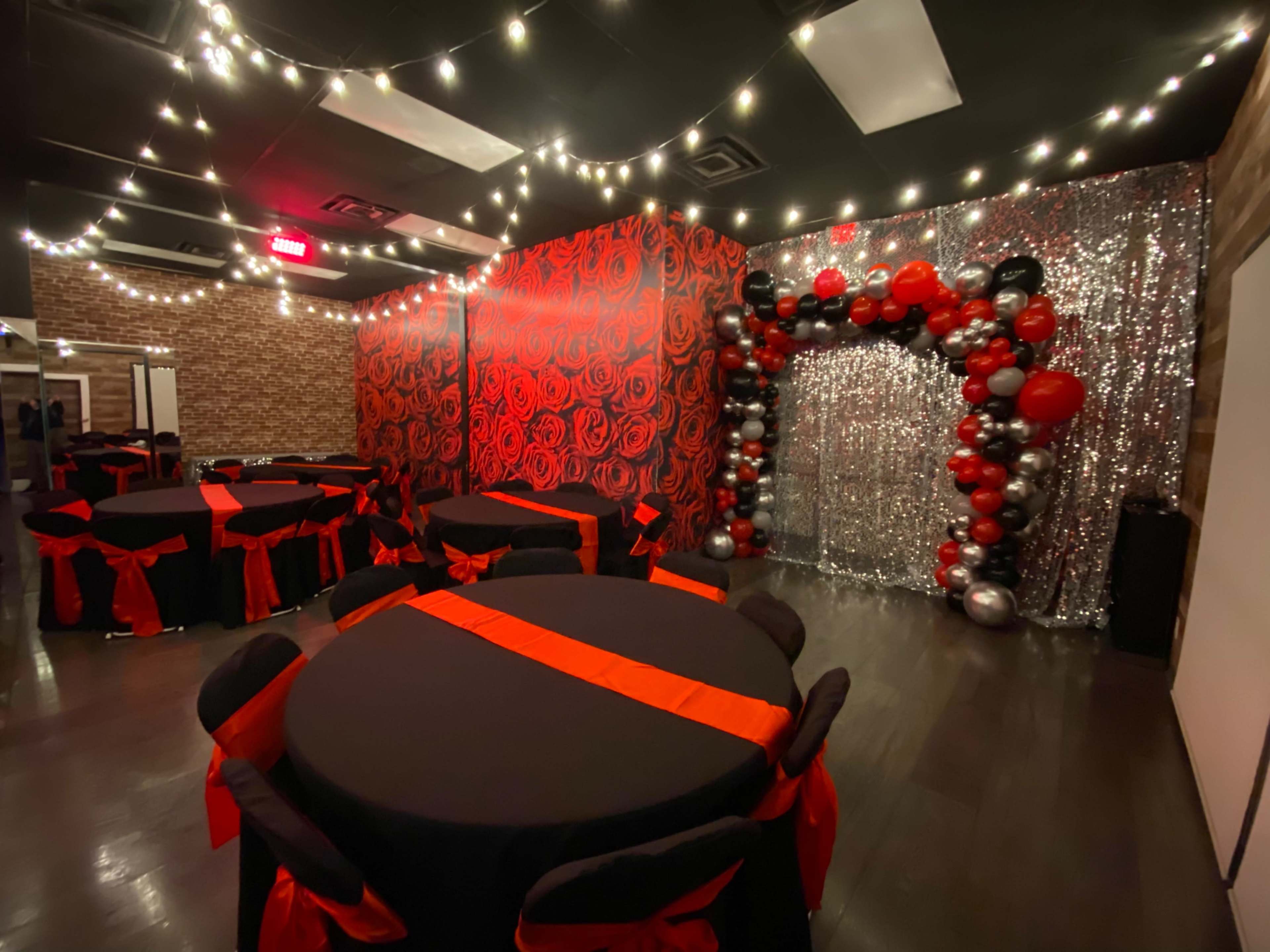 A decorated event space with black tables featuring red sashes, a wall adorned with red roses, and an arch made of silver and black balloons against a shimmering backdrop.