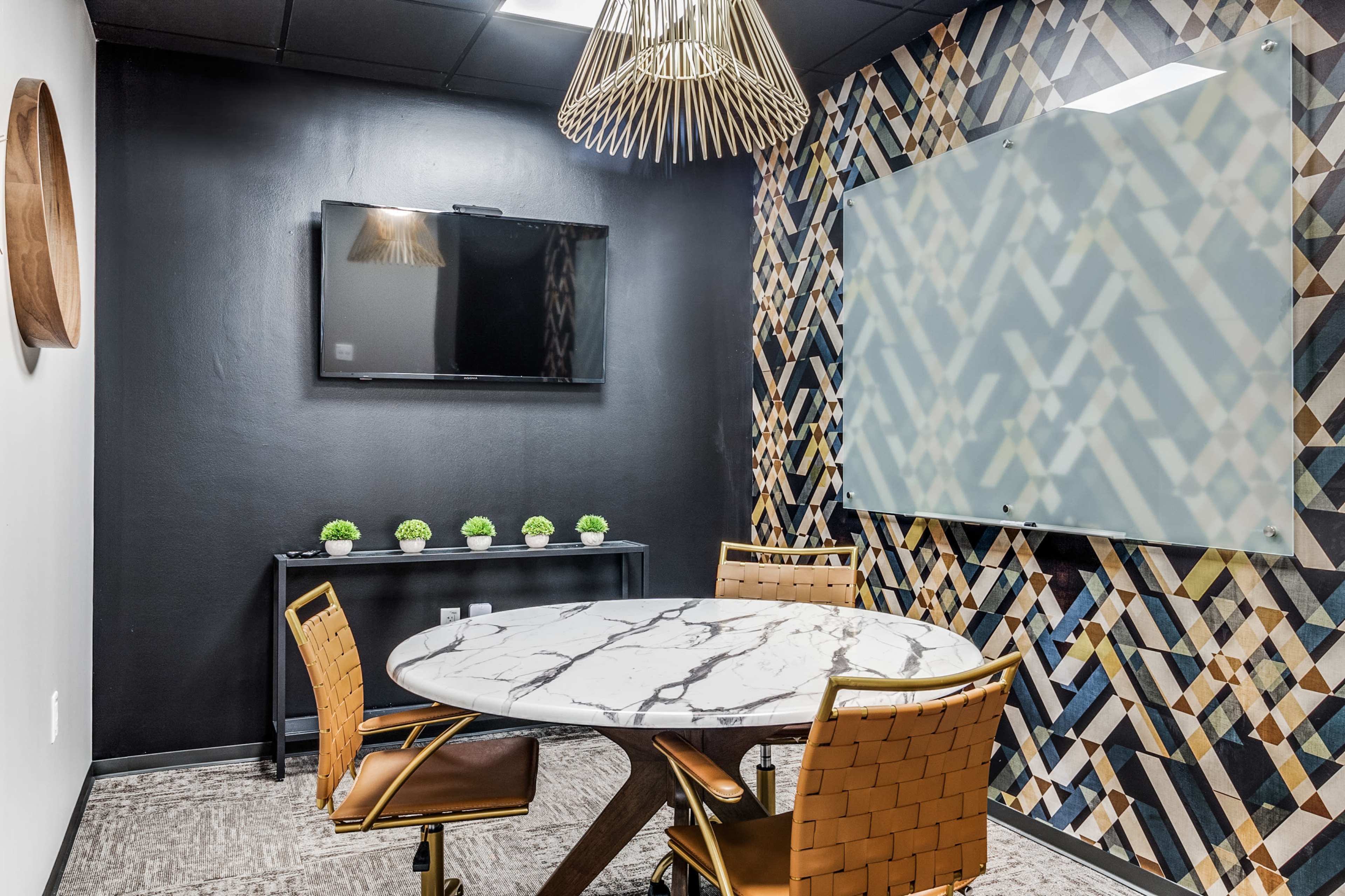 A modern meeting room features a round marble table with gold chairs, a flat-screen TV on the wall, and a decorative patterned backdrop.