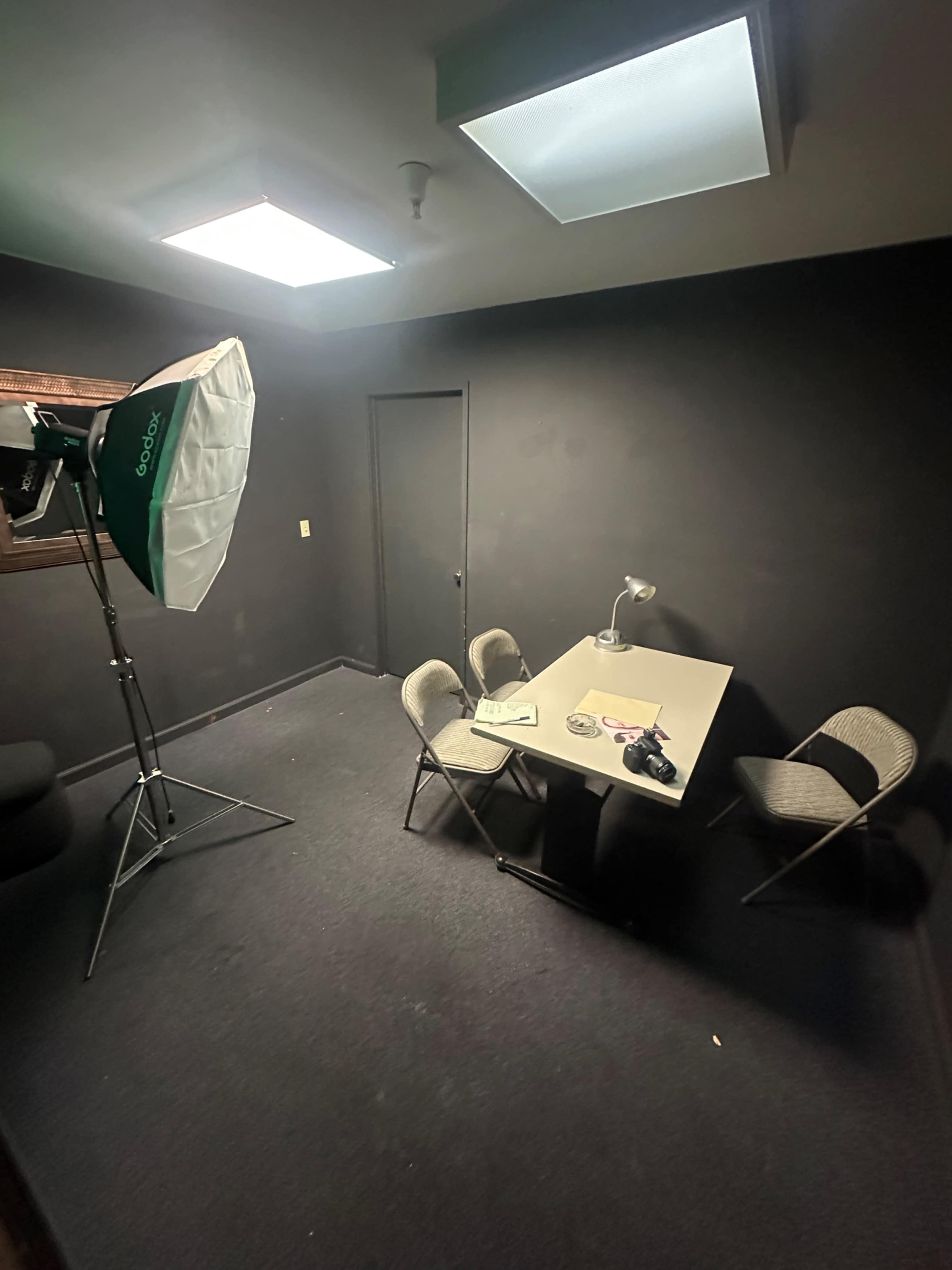 The image shows a dimly lit room with black walls, a table and three chairs, and a photography light set up in one corner.