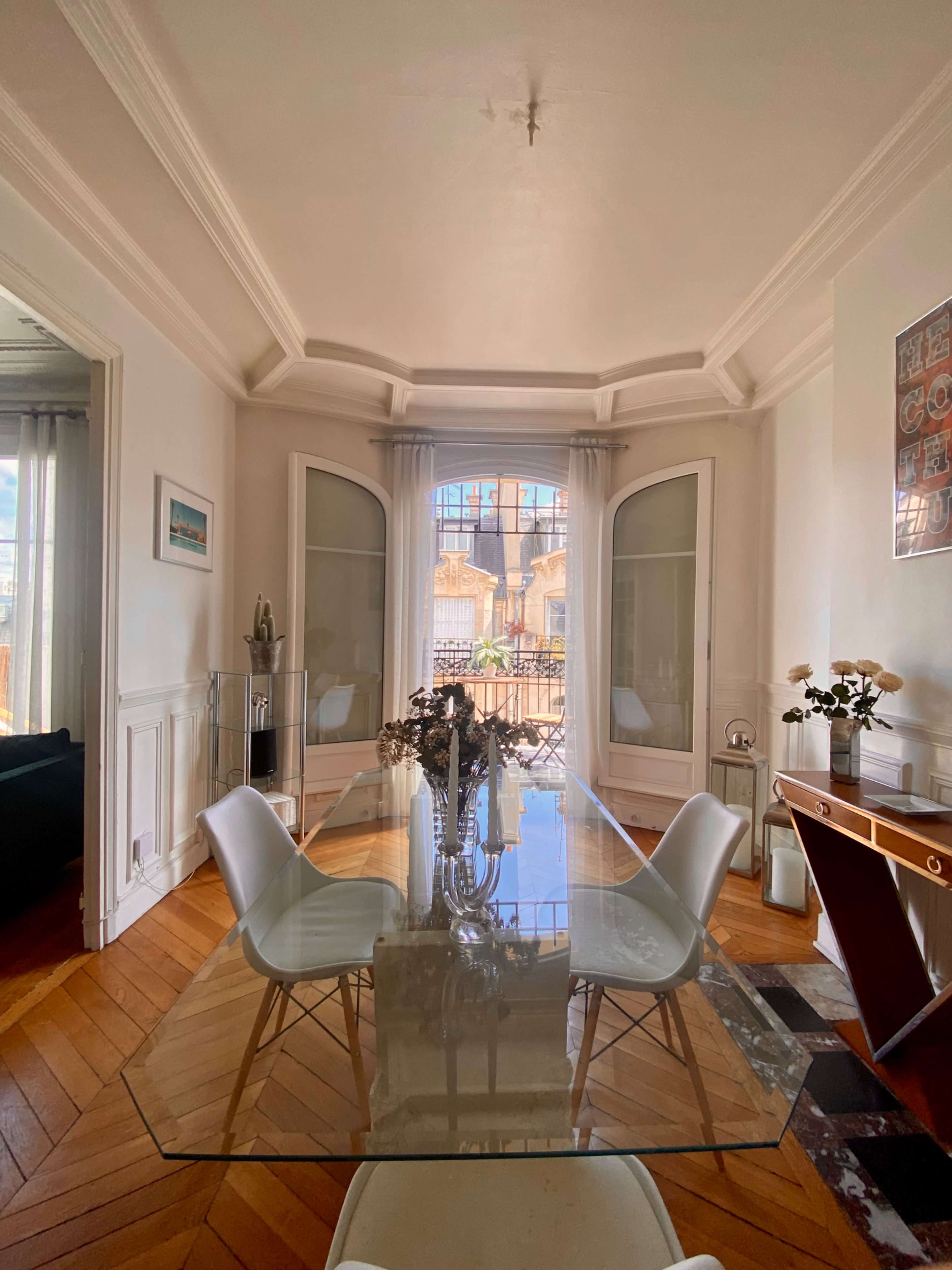 A glass dining table is set in a brightly lit room with large windows and wooden flooring.