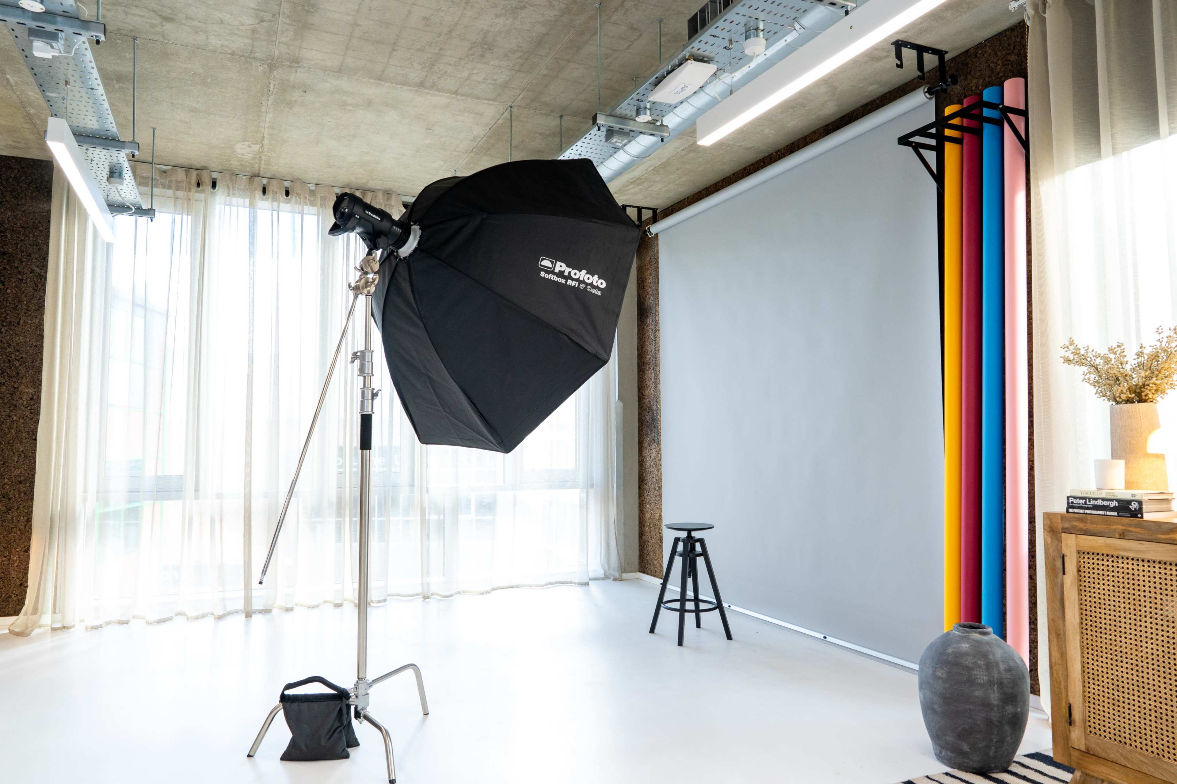 The image shows a photography studio setup with a large softbox light, a background with colorful drapes, and a stool positioned nearby.