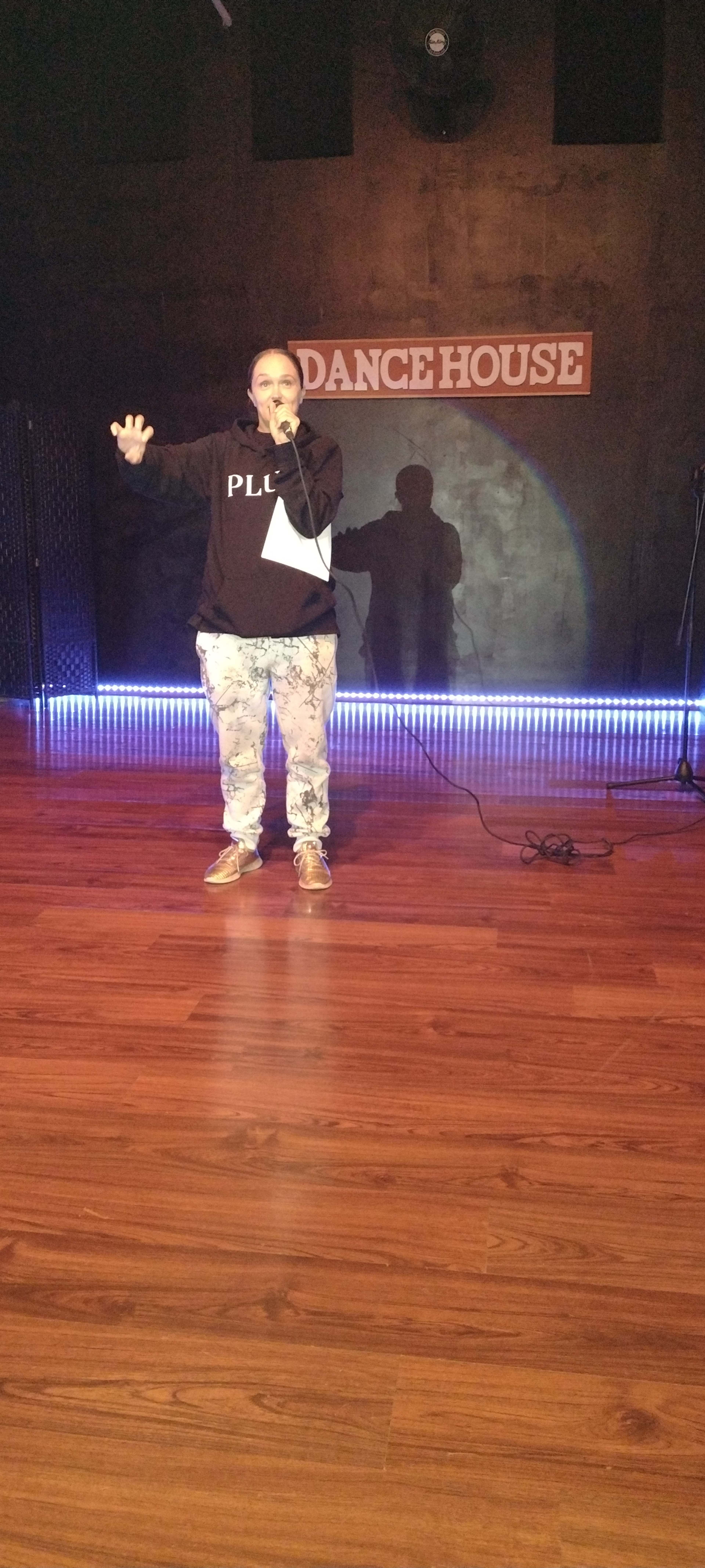 A person stands on a wooden stage in a performance space, holding a microphone and facing an audience with a shadow cast on the wall behind them.