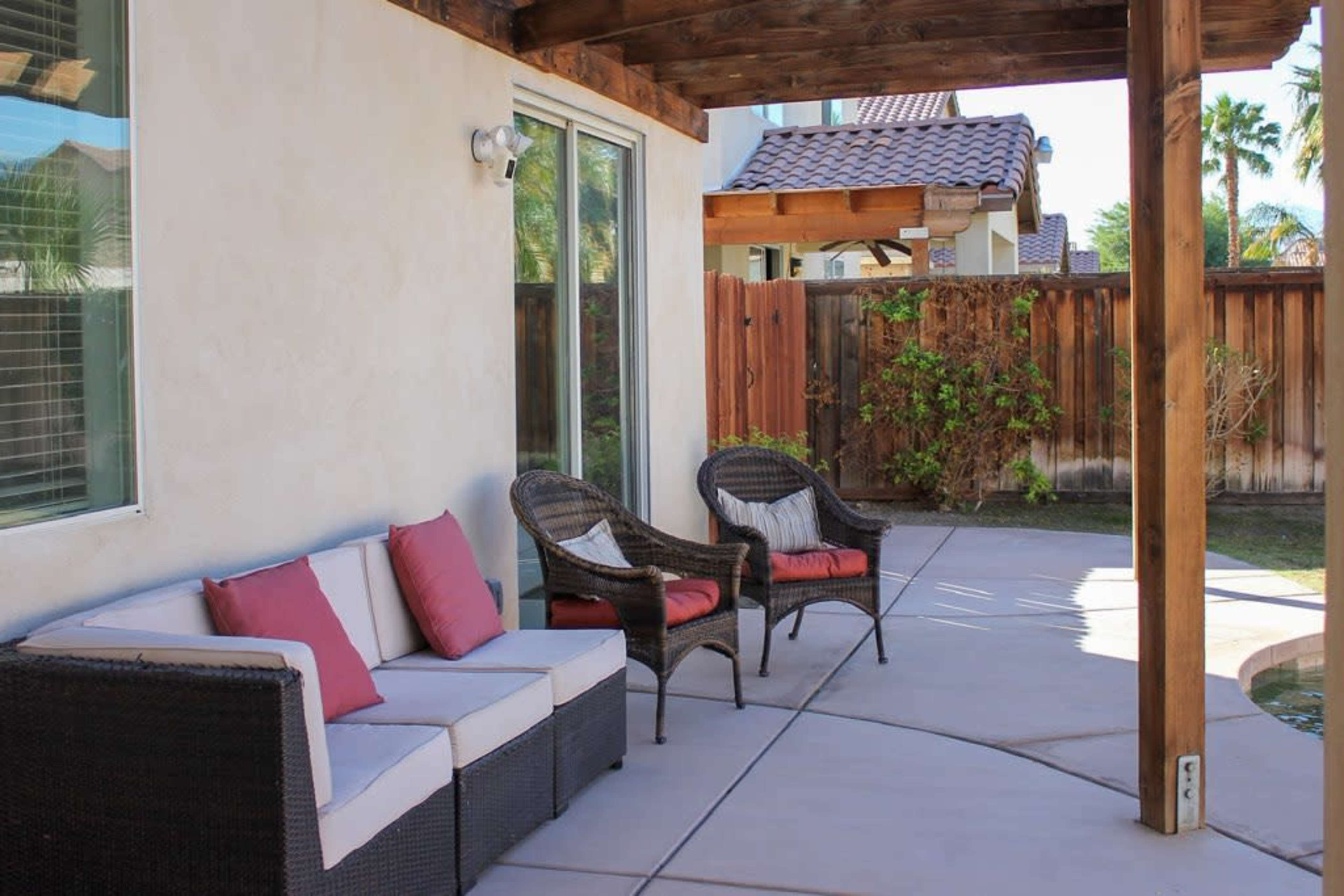 A patio area featuring a couch and two chairs with cushions, situated beside a swimming pool and surrounded by a wooden fence and greenery.