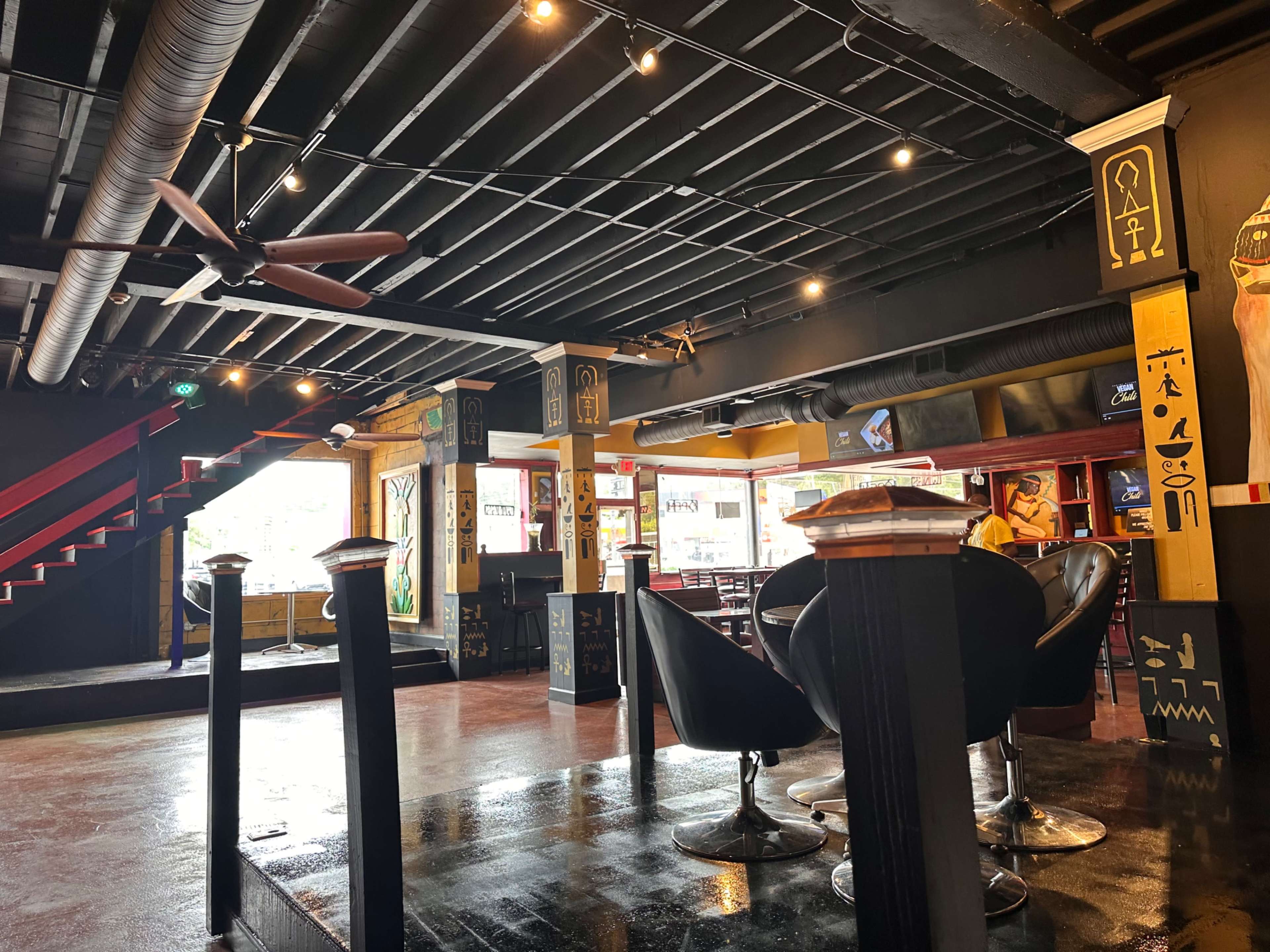 The interior of a restaurant features black walls, wooden beams, and a staircase leading to an upper level, with chairs arranged around tables.