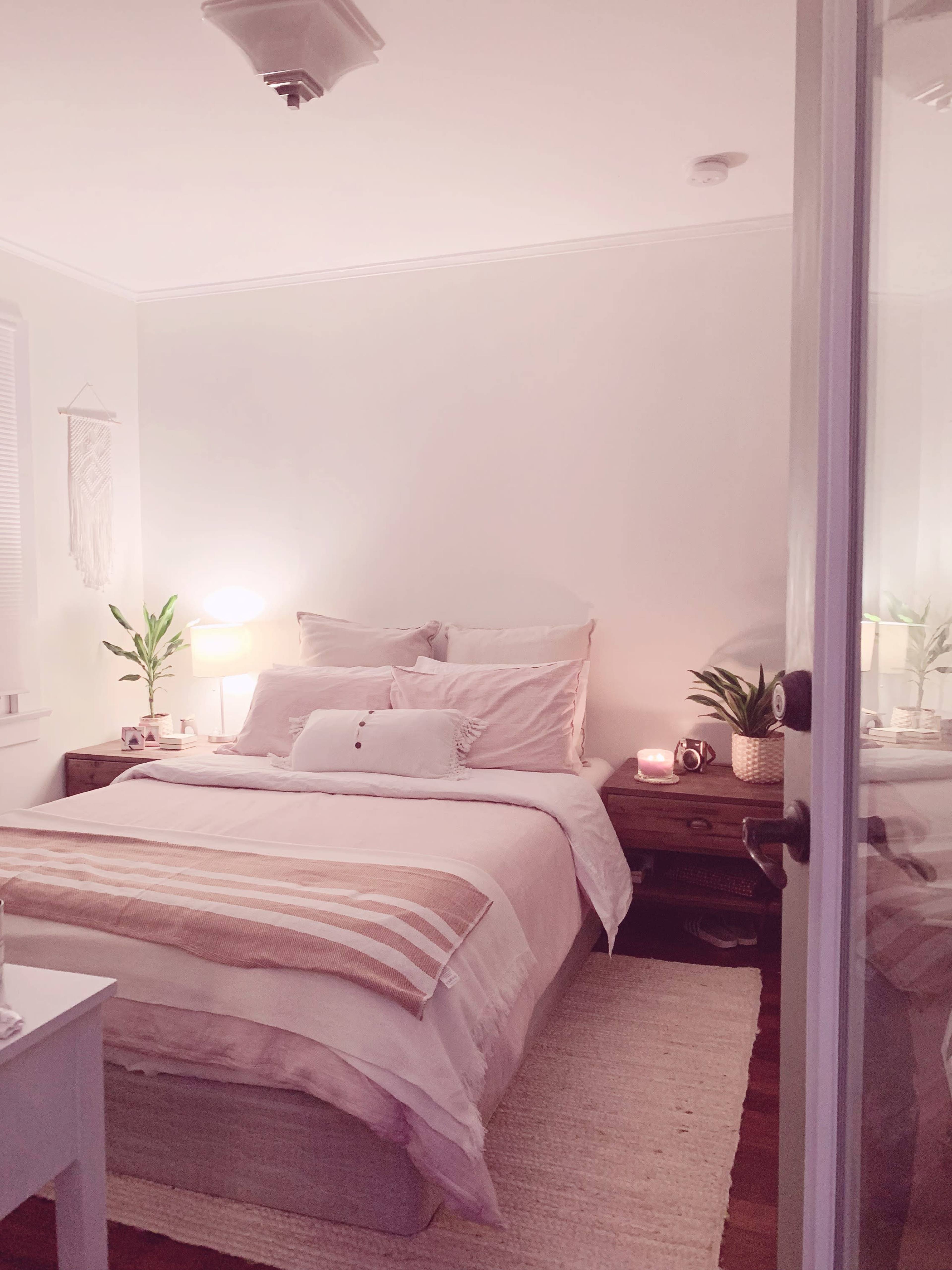 The image shows a cozy bedroom with a neatly made bed, bedside tables with lamps, and decorative plants on the furniture.