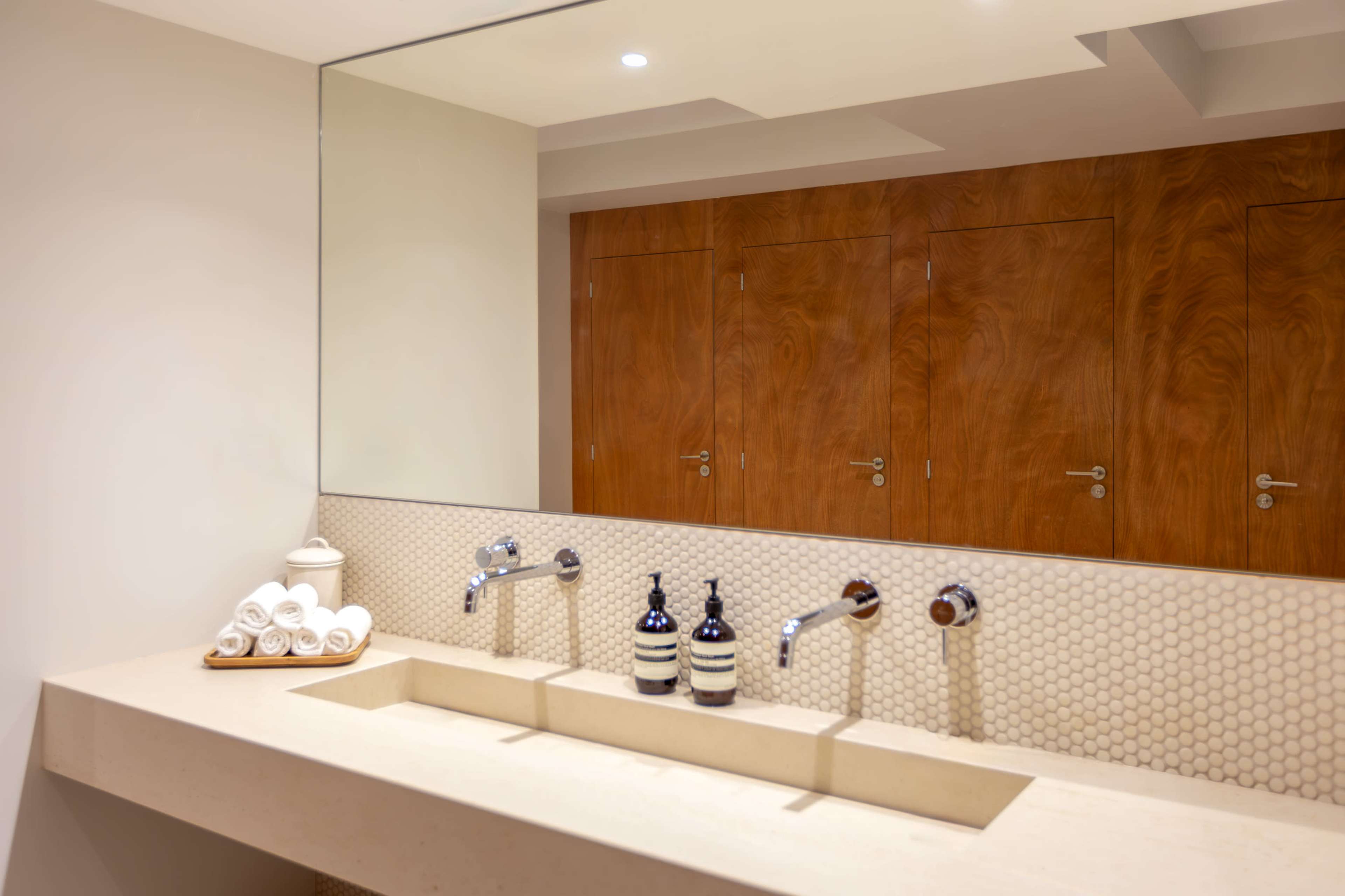 A modern bathroom features a long, dual sink counter with stylish faucets and neatly arranged towels, while wooden doors serve as the backdrop.