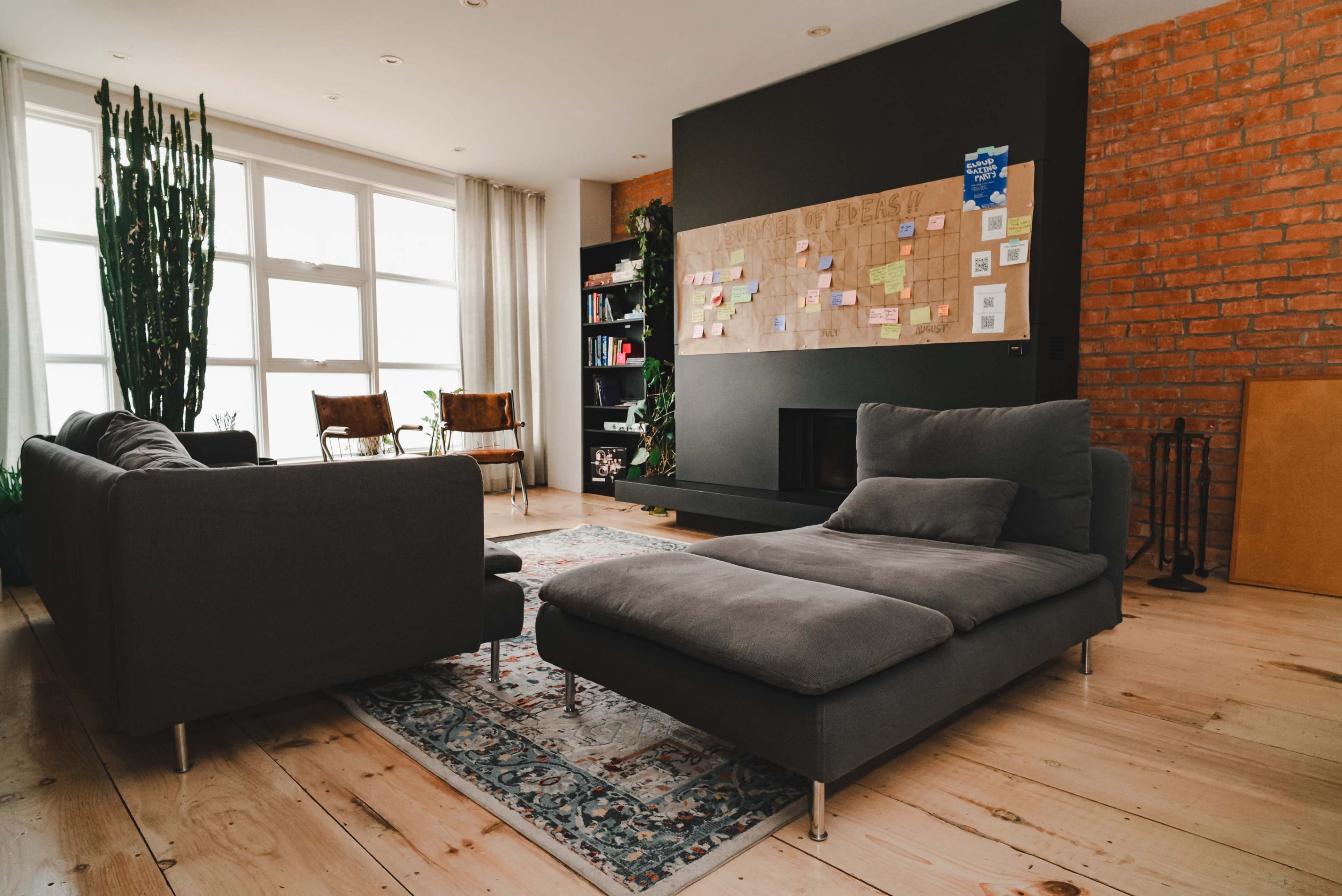 The scene features a modern living room with a sectional sofa, a large bulletin board on the wall, and potted plants near a window, complemented by wooden flooring and exposed brick accents.