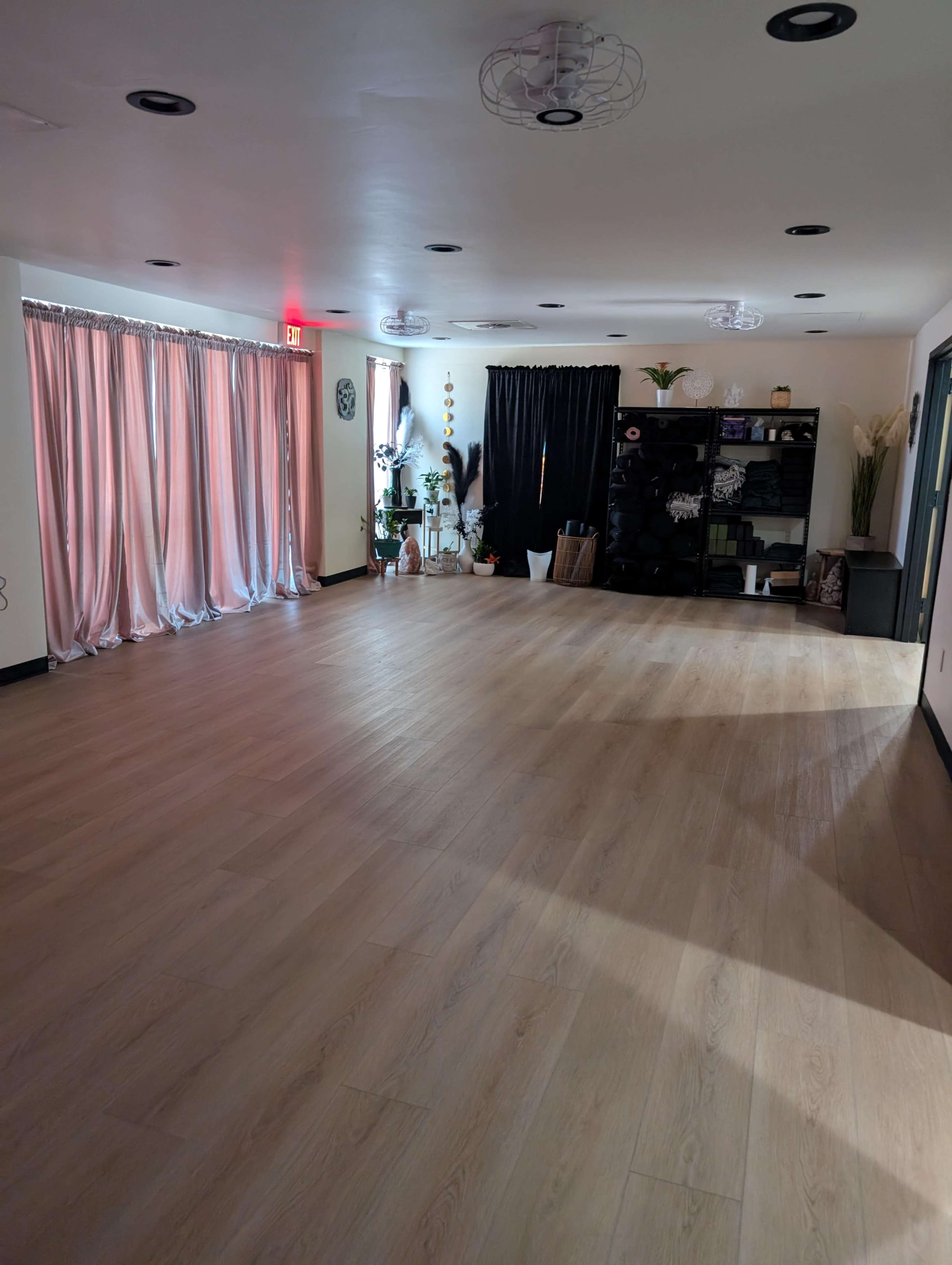 The image shows an empty room with wooden flooring, large windows covered by sheer curtains, and a black storage unit along one wall.