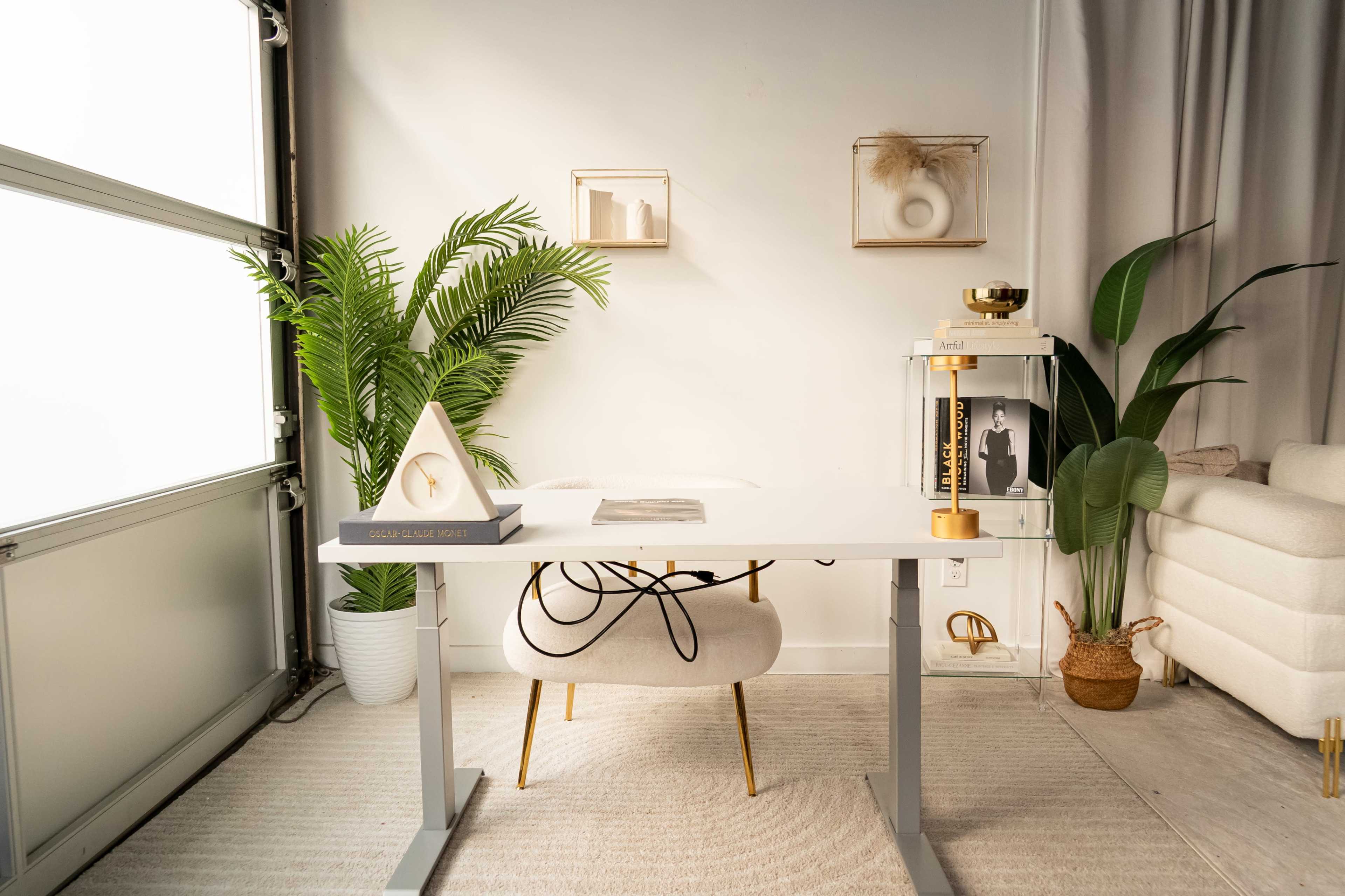 A modern office space features a minimalist white desk, a stylish chair, decorative plants, and framed art on the walls.