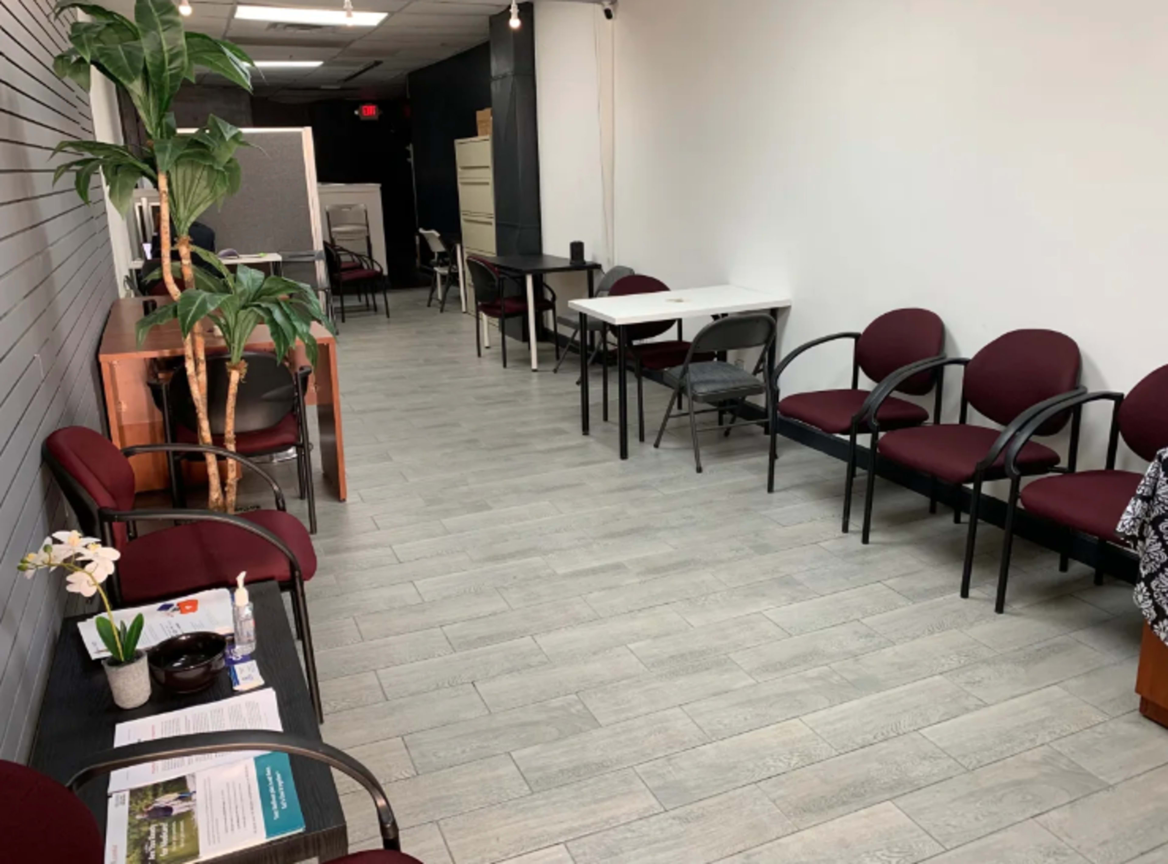 A clean, spacious waiting area with several maroon chairs arranged along a light gray floor, featuring a small table and a few green plants.