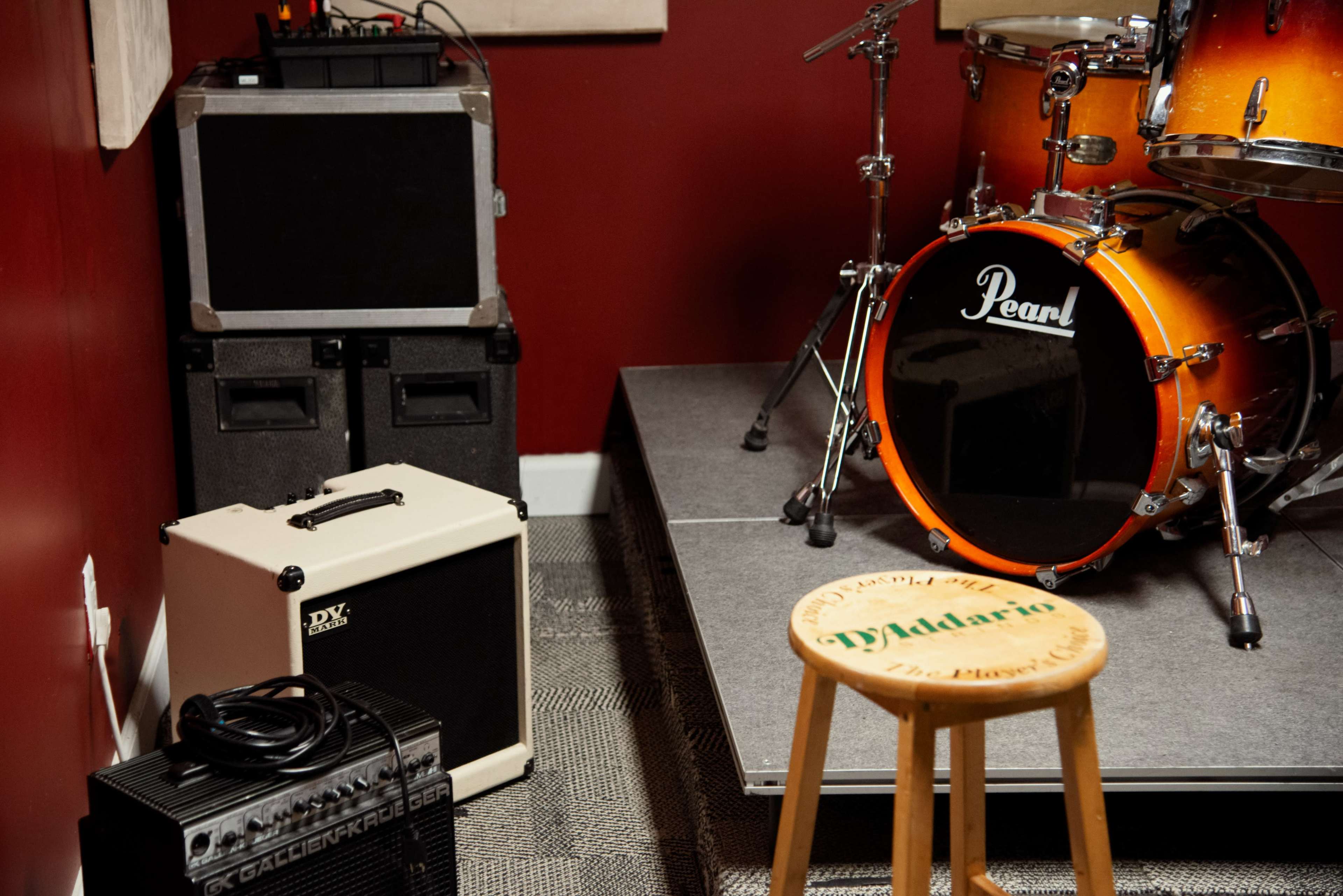 The image shows a music practice space featuring a Pearl drum set, an amplifier, a small stool, and several equipment cases against a red wall.