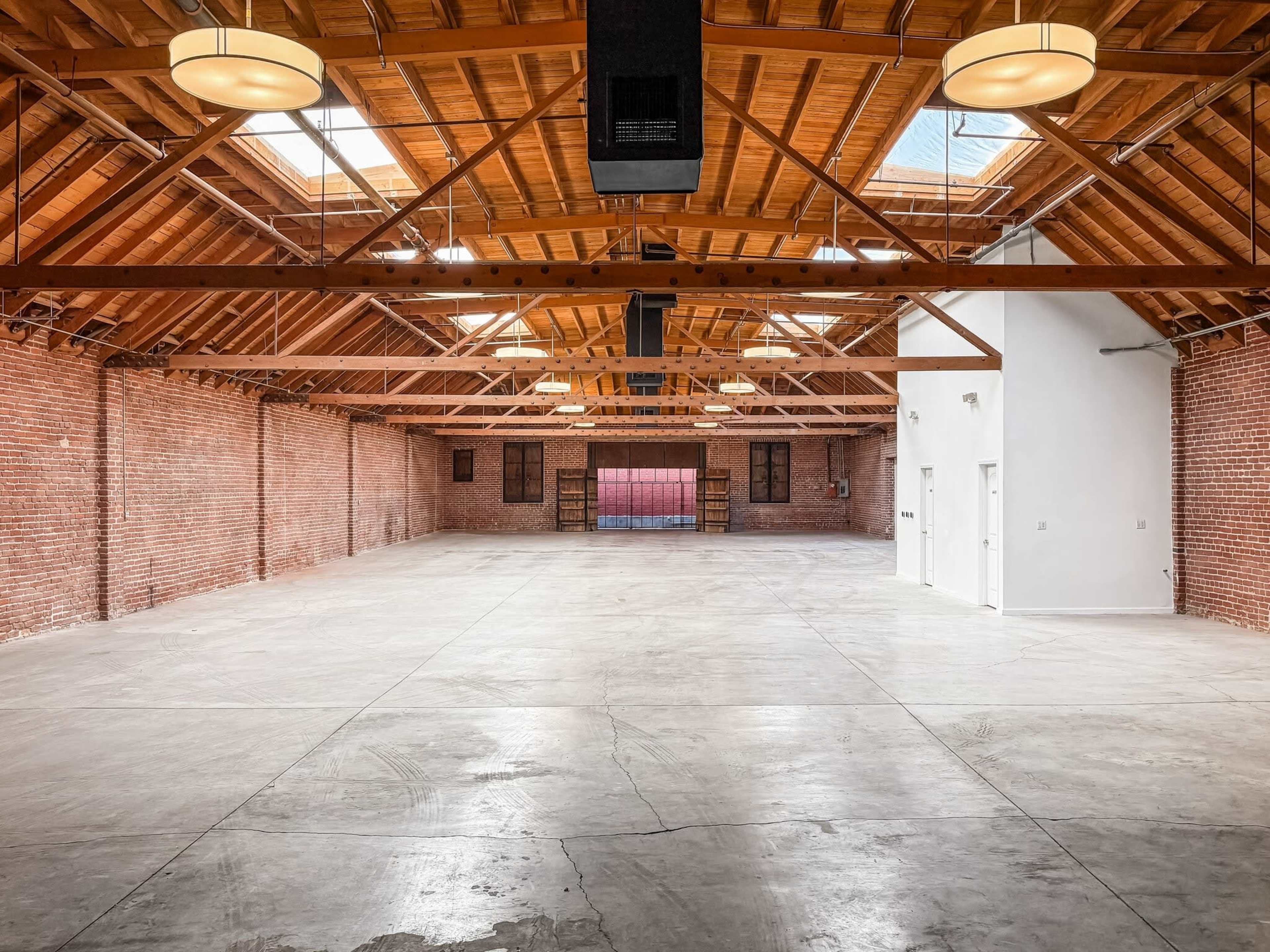 The image shows a spacious, empty warehouse with exposed brick walls and a wooden beam ceiling featuring skylights.