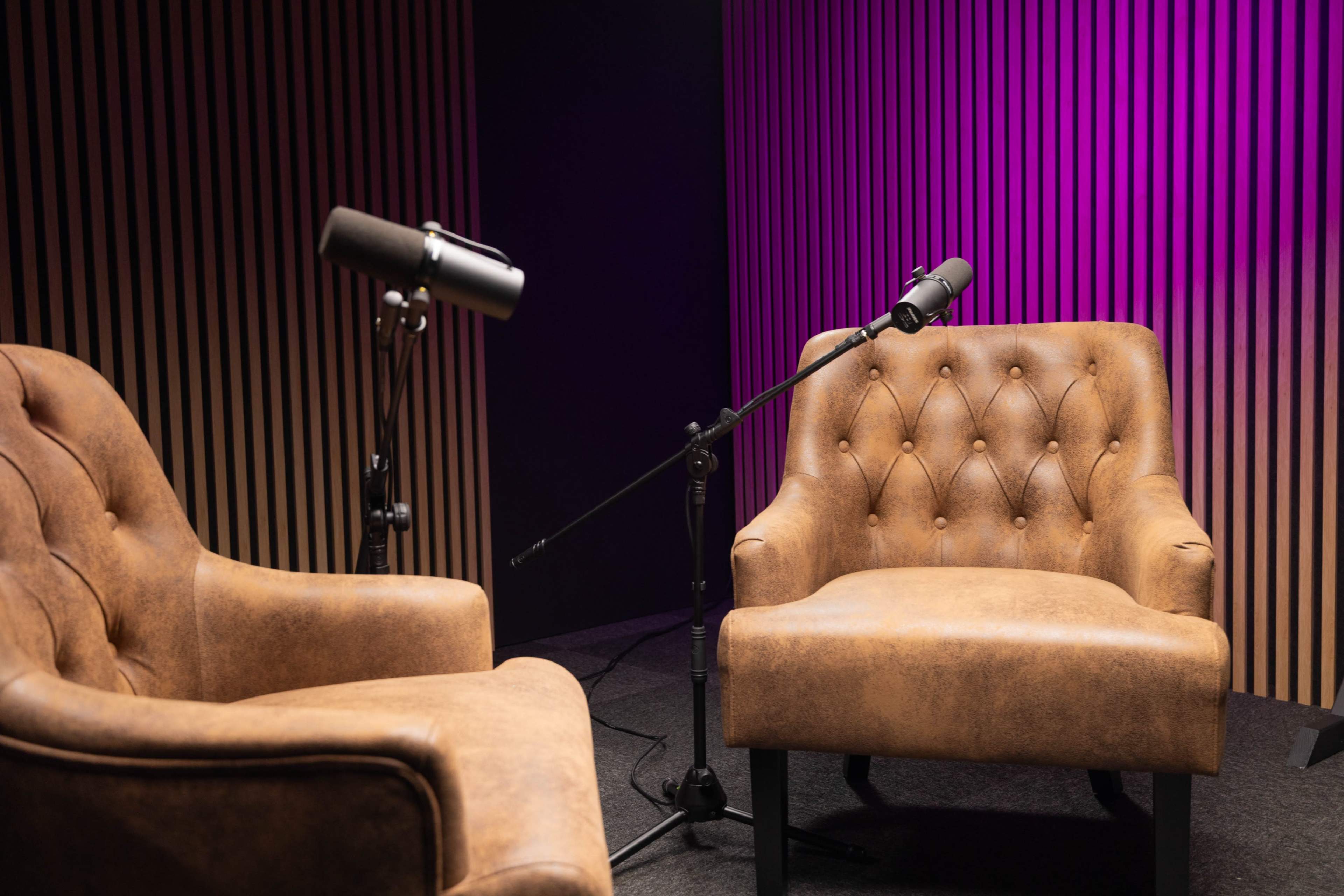 The image shows two brown leather armchairs positioned opposite each other, each with a microphone mounted beside them against a backdrop of vertical wooden panels illuminated in purple light.