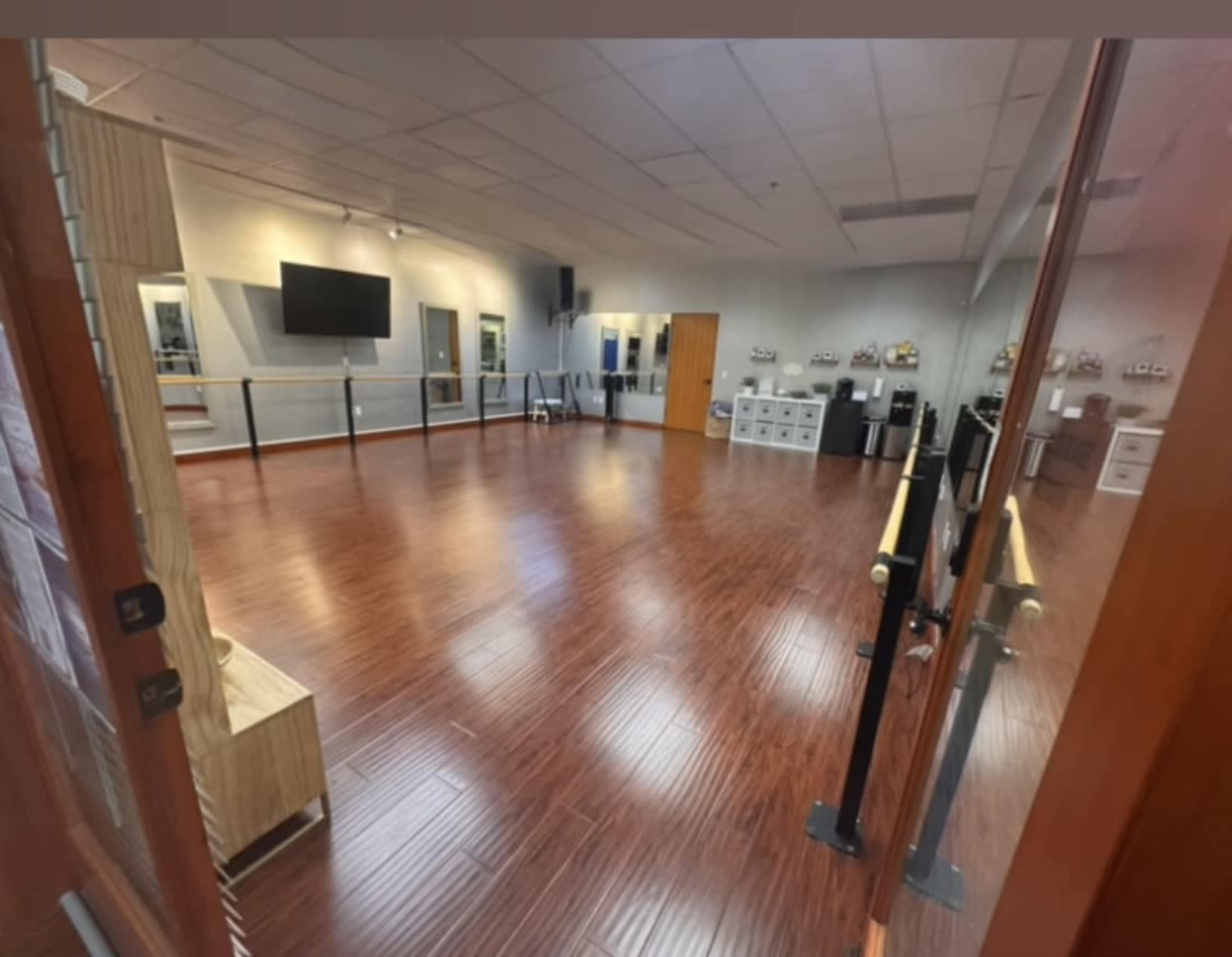 The image shows an empty dance studio with wooden flooring, mirrors on the wall, and ballet barres along the sides.
